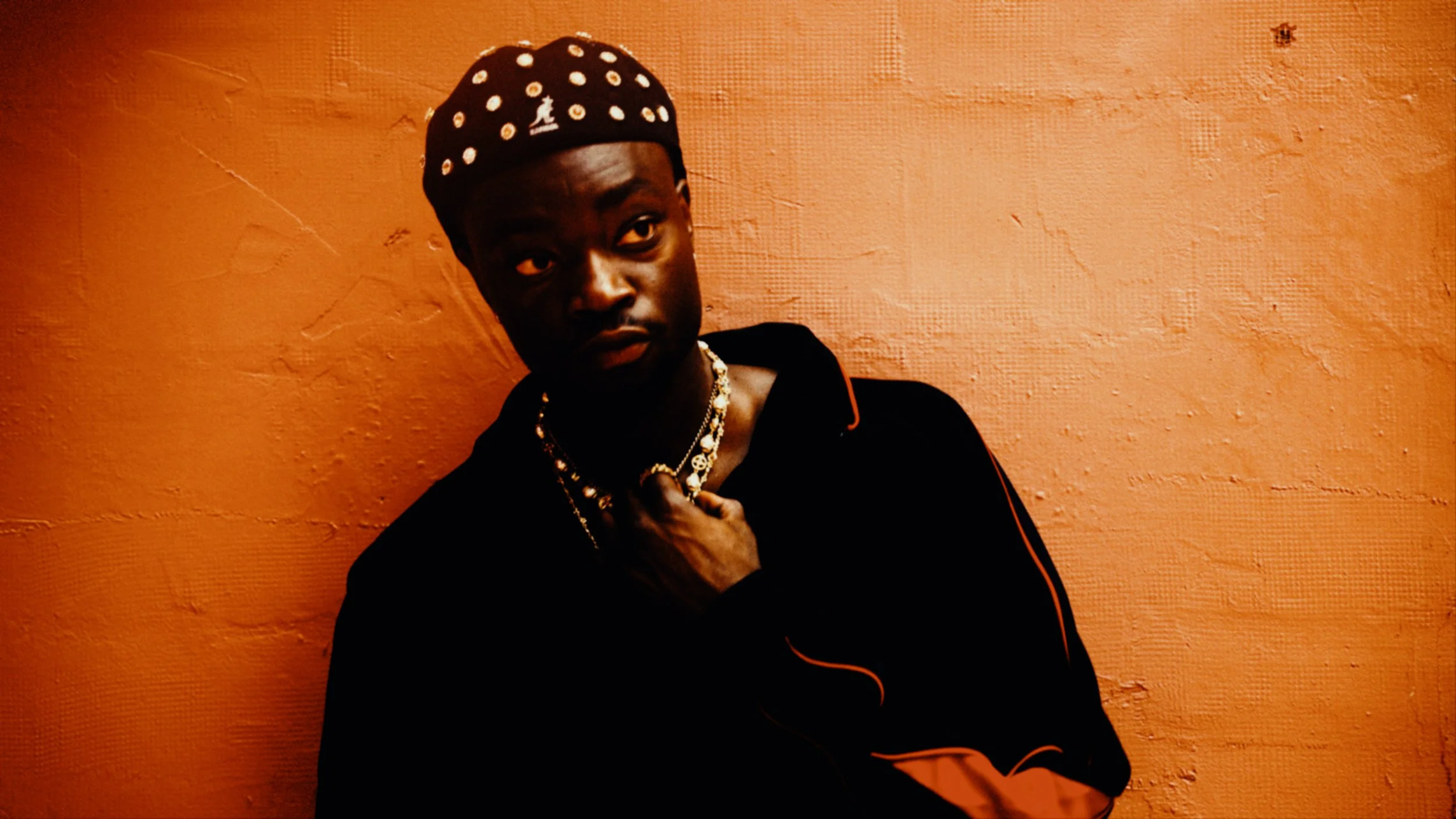 A young man wearing a black hoodie with orange accents, a patterned black and white hat, and gold jewelry, standing against an orange textured wall.