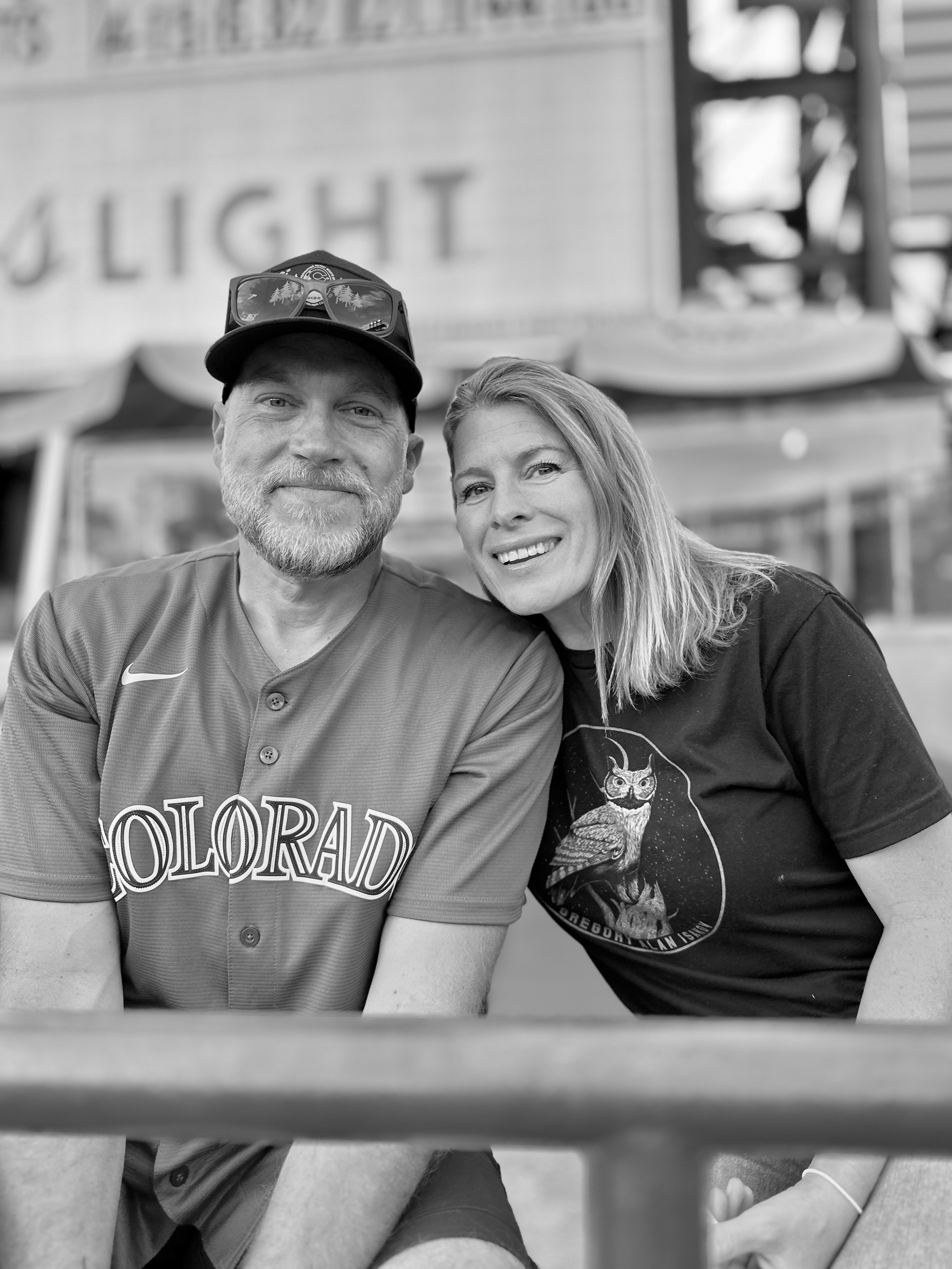 A smiling man and woman sitting closely together at an outdoor location, with a blurred background and partial view of a sign.