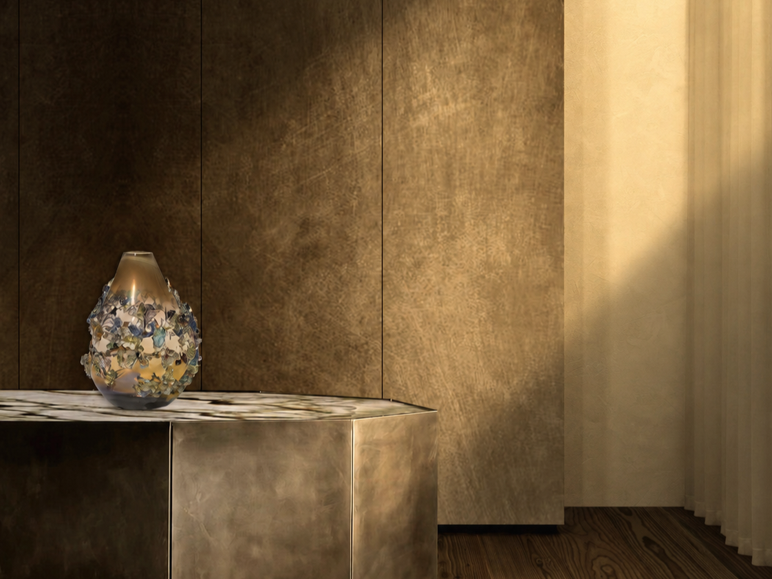 A decorative vase with floral patterns on a metallic surface, with a golden textured wall behind and a curtain on the right side.