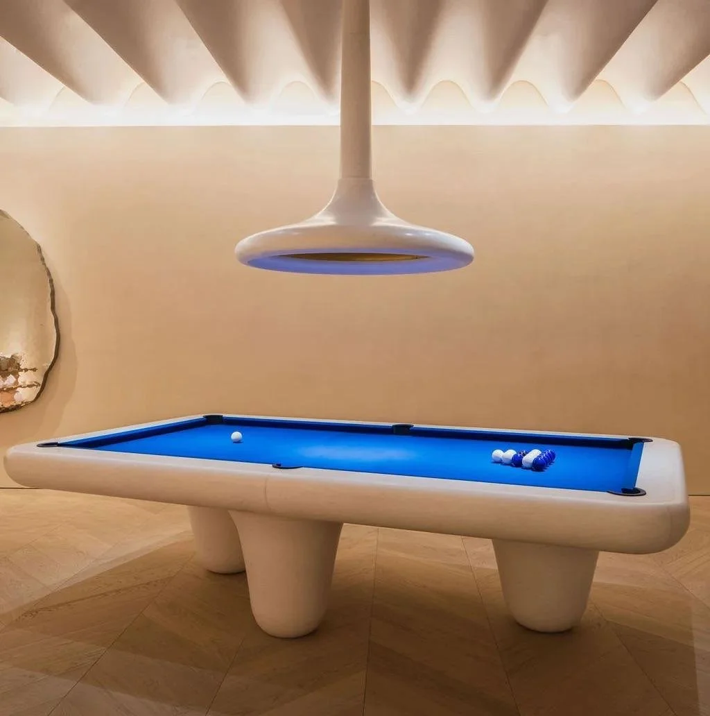 A modern, curved pool table with blue felt, white balls, a rack of striped balls, and a single white cue ball, under a large, futuristic white pendant light in a minimalistic room.