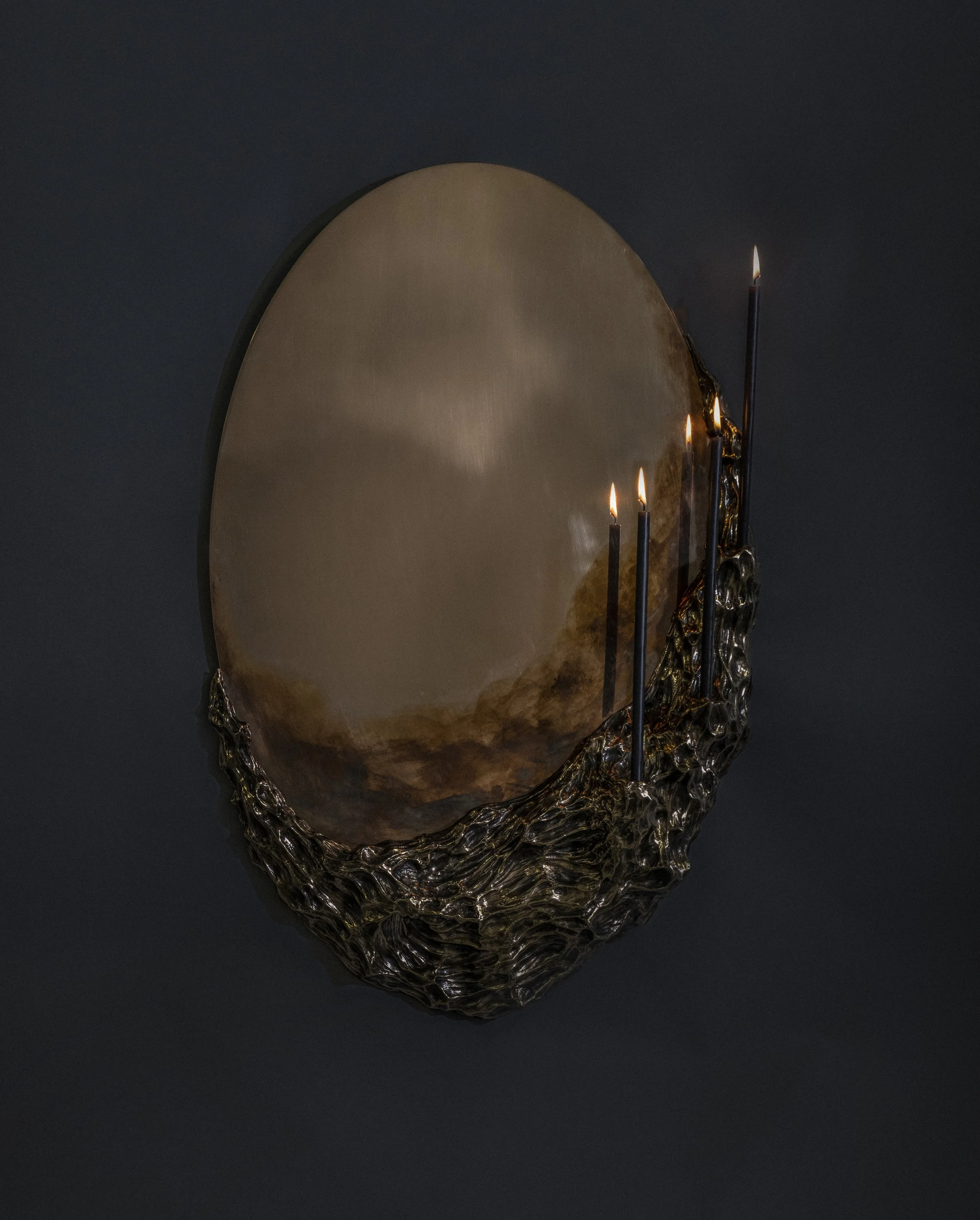 A decorative mirror with a textured, dark metallic frame and three lit candle sticks attached to the side, against a dark background.