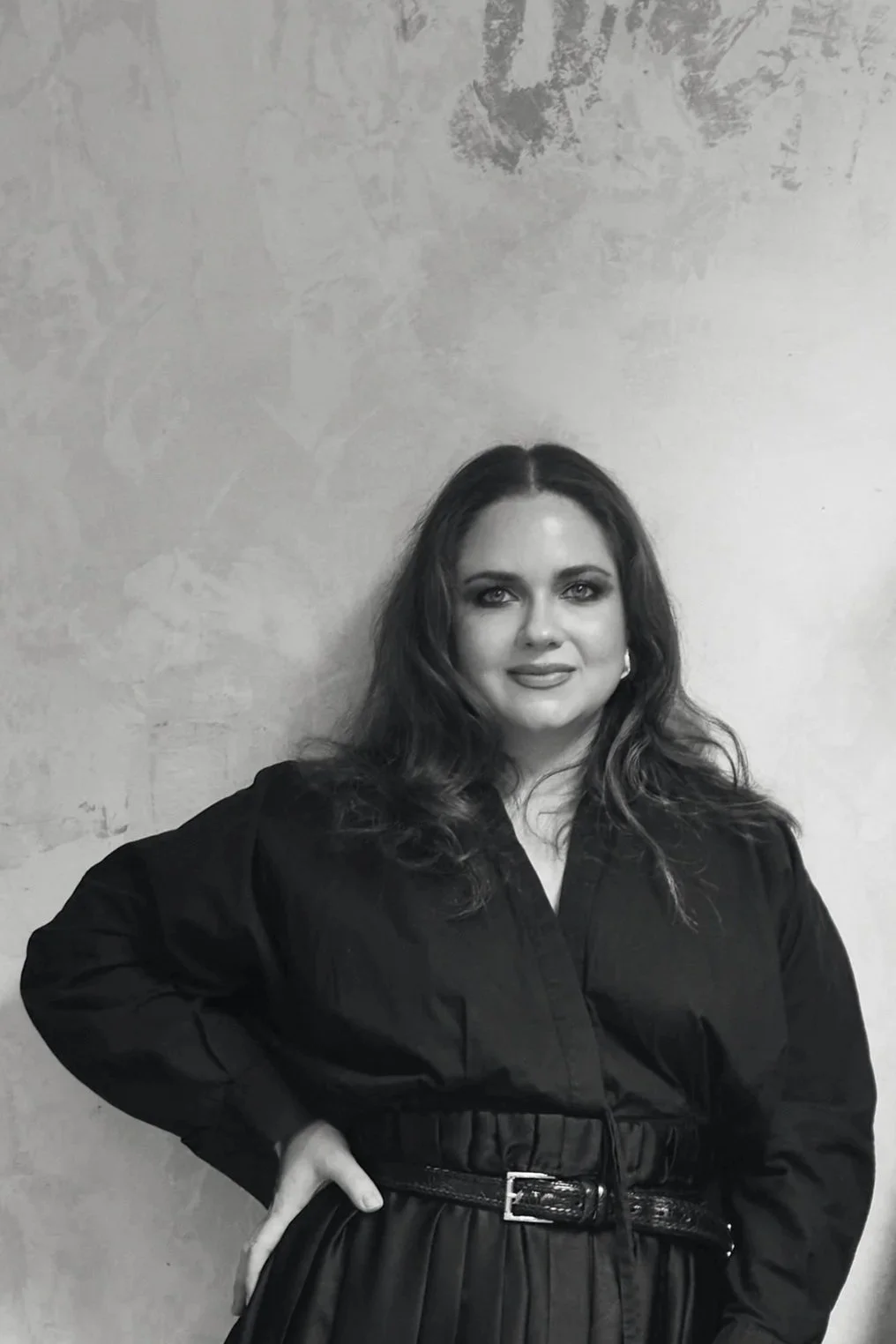 Black and white portrait of a woman with long wavy hair, posing with her head resting on her hand, wearing a dark blouse, against a plain background.