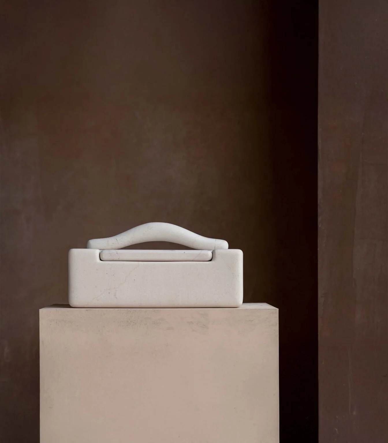 A white, minimalist handbag or clutch placed on a beige pedestal against a dark background.