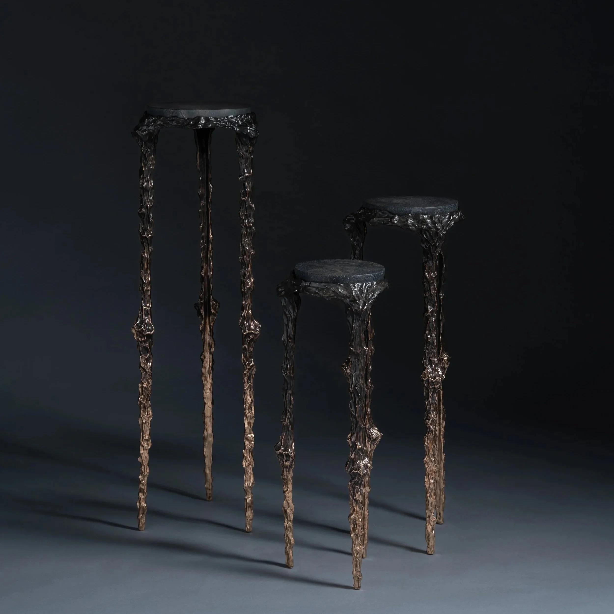 Three tall, dark, ornate metal stools with textured, irregular legs and small, rounded seats on a dark background.