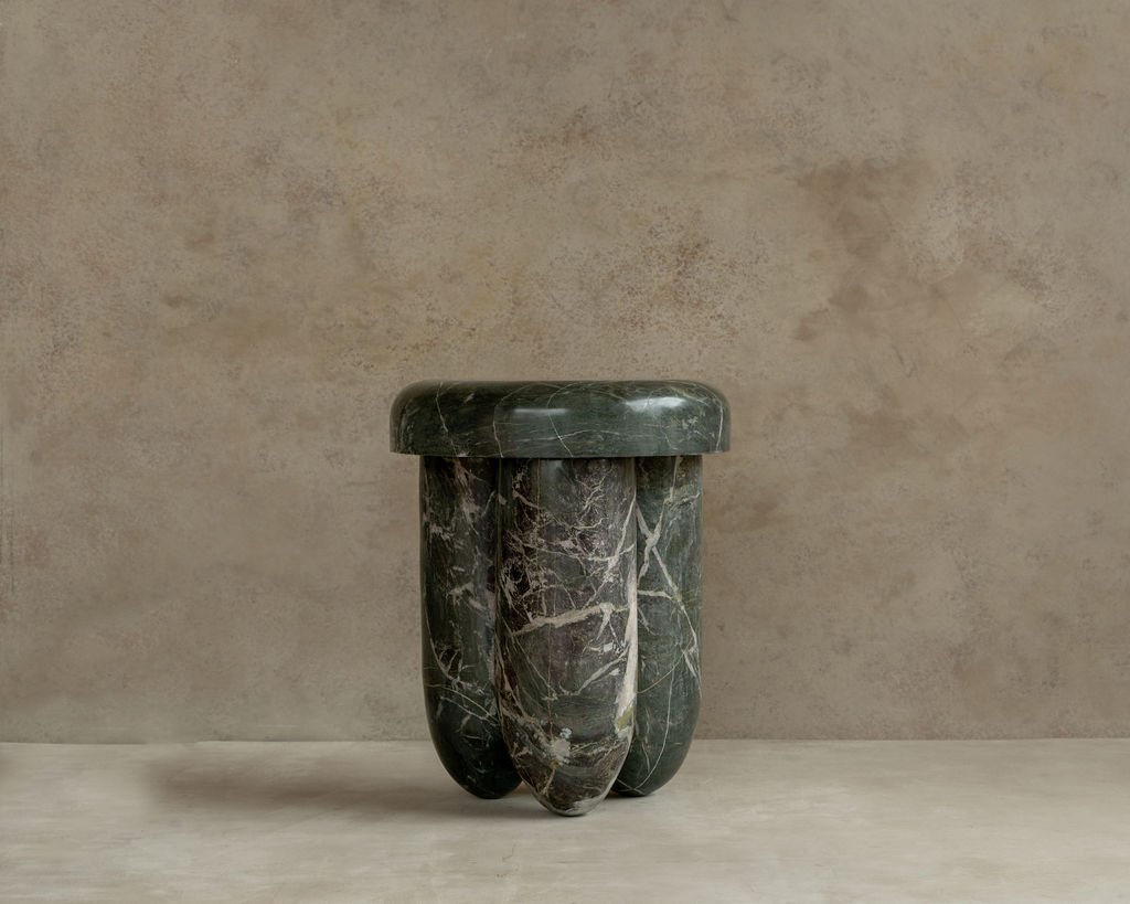 A dark green marble table with a rounded top and three thick, rounded legs, set against a beige textured wall.