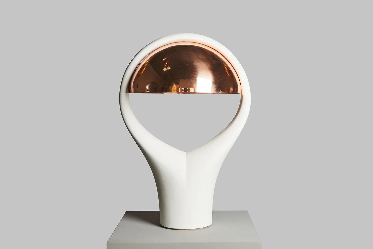Modern sculpture with a white base and a half-sphere copper-colored top, displayed on a white pedestal against a gray background.