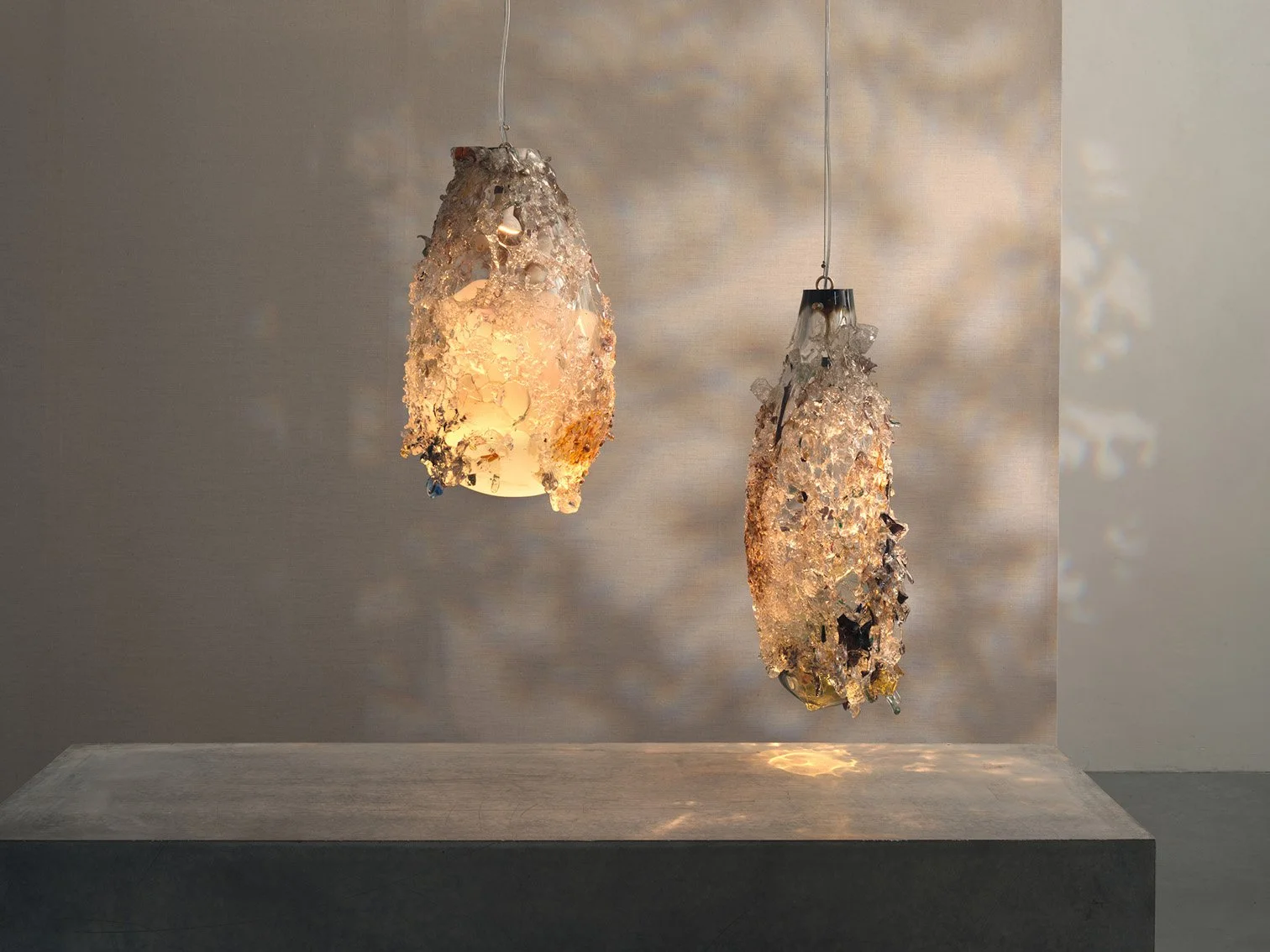 Two hanging light fixtures covered with melted glass or plastic shards, casting intricate shadows on the wall below, against a neutral background.