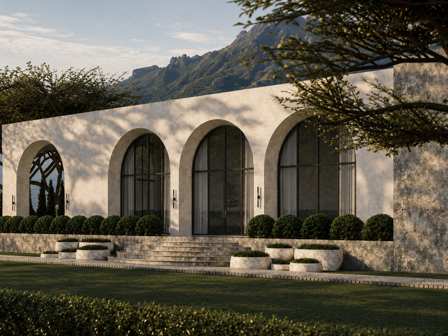 Modern building with large arched glass doors and windows, surrounded by bushes and greenery, with mountains in the background.