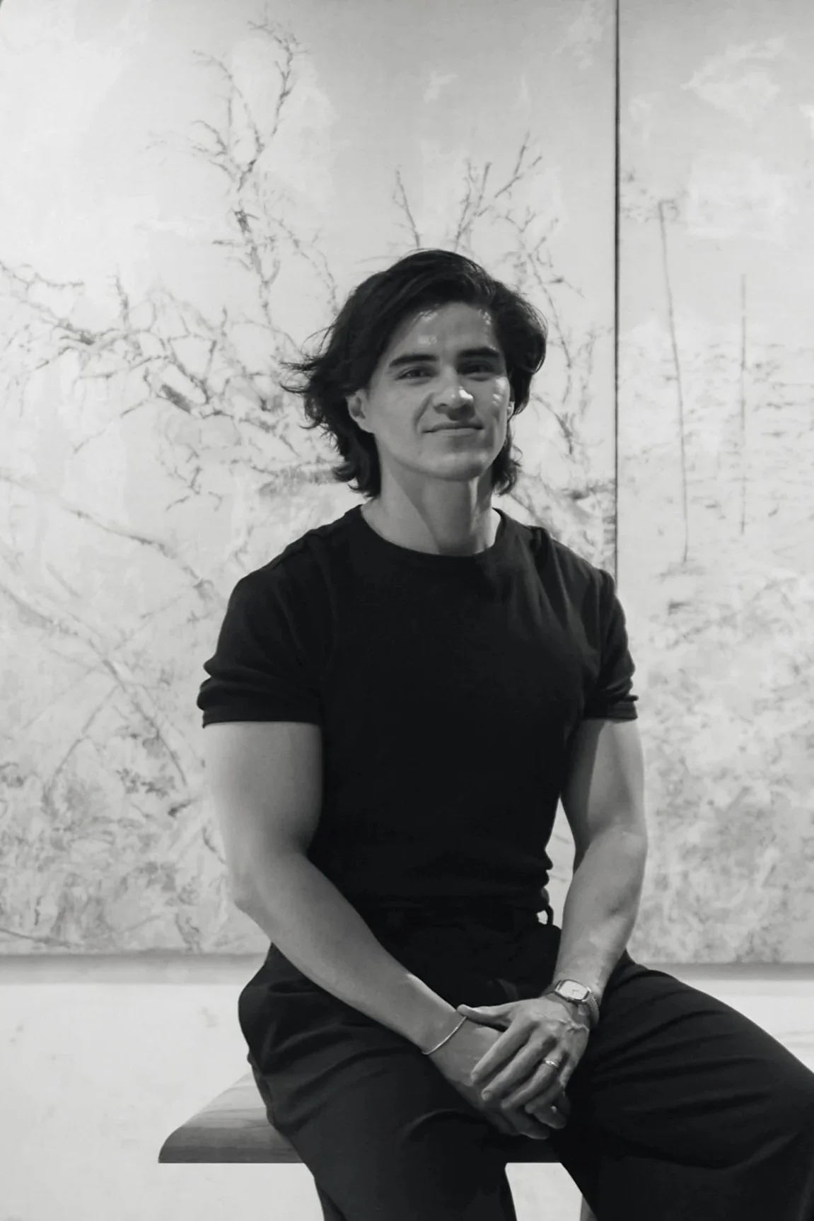 A young man with dark, styled hair, wearing a plain white t-shirt and dark pants, sitting on a marble block against a plain gray wall, looking thoughtfully to the side in black and white.