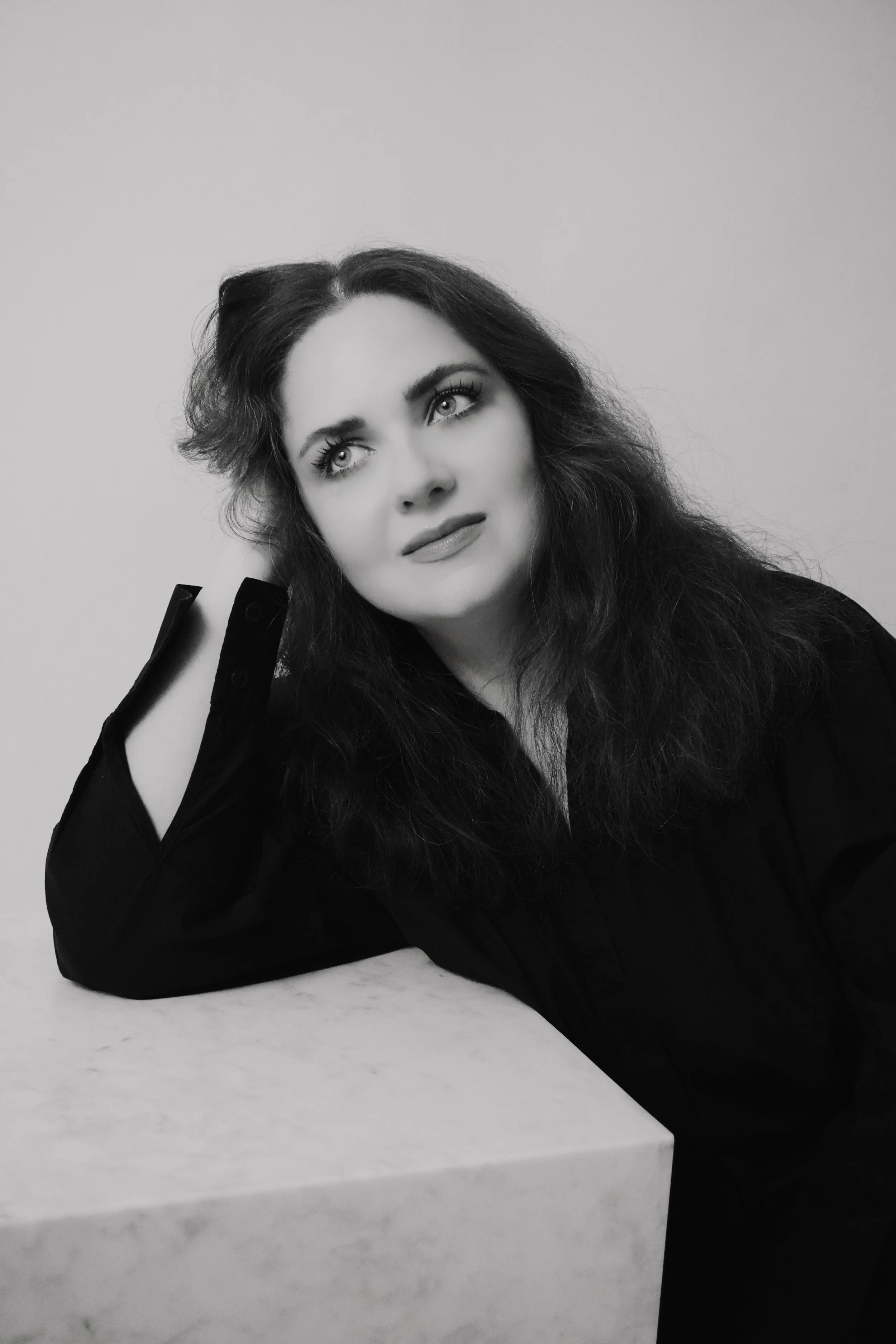 Black and white portrait of a woman with long wavy hair, posing with her head resting on her hand, wearing a dark blouse, against a plain background.