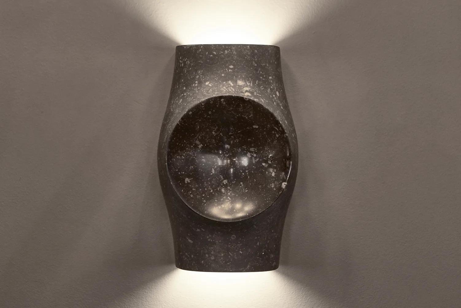 Modern wall-mounted light fixture with a rounded, oval shape, illuminating the wall above and below, with a speckled dark surface.