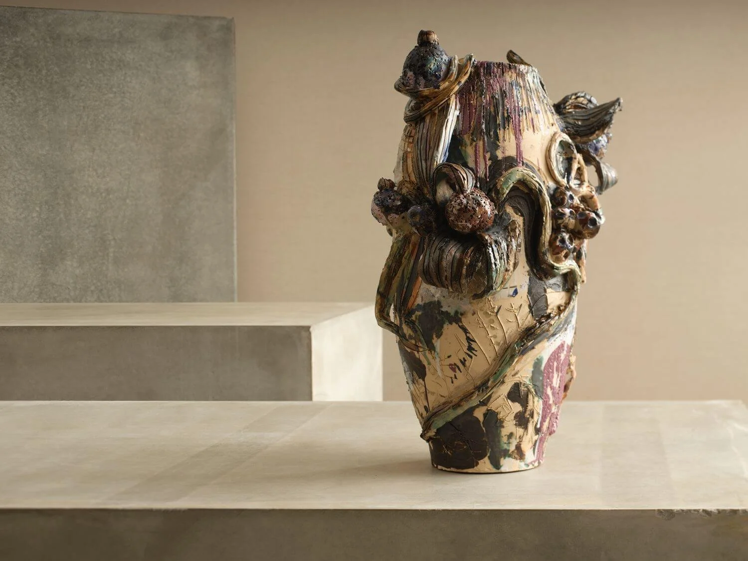 Artistic ceramic vase with textured, colorful, abstract figures and patterns, displayed on a beige pedestal against a plain background.