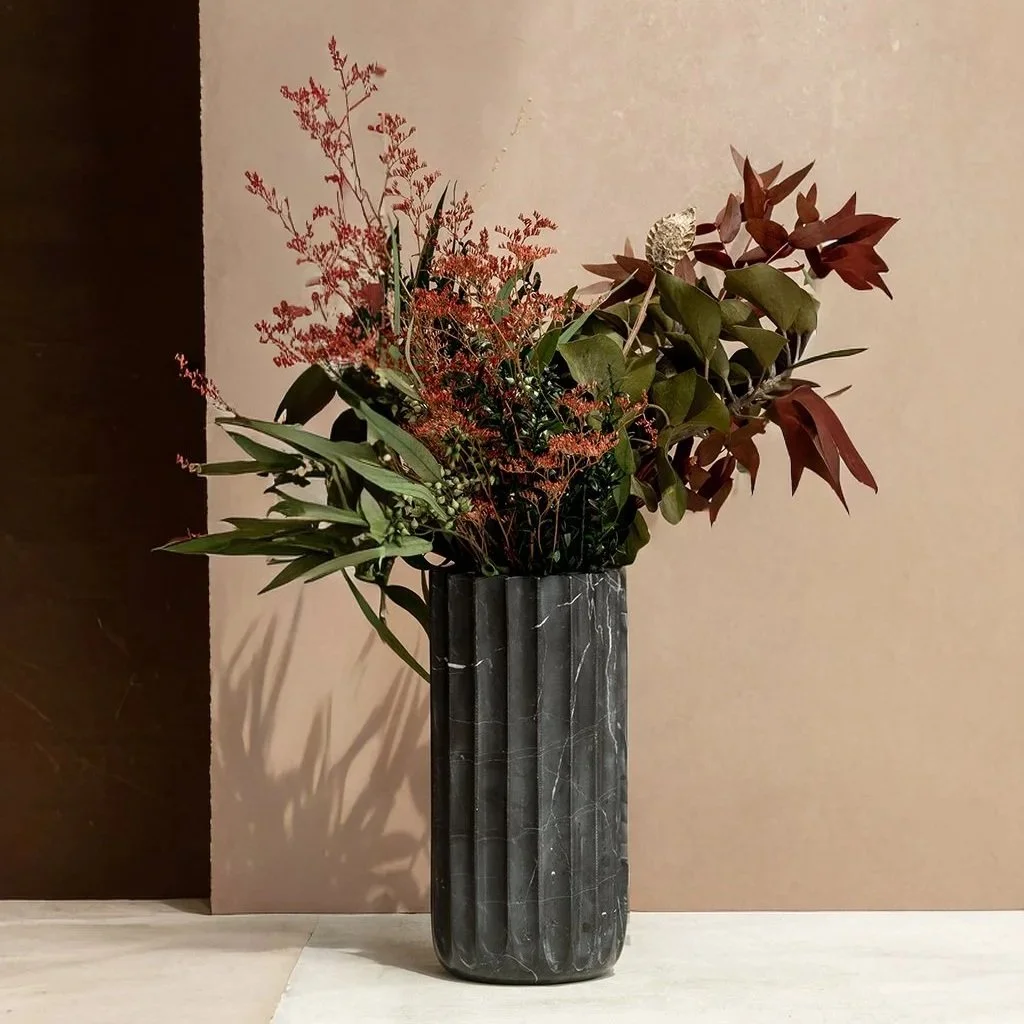 A tall black marble vase with various types of green and reddish-brown leaves and dried flowers.