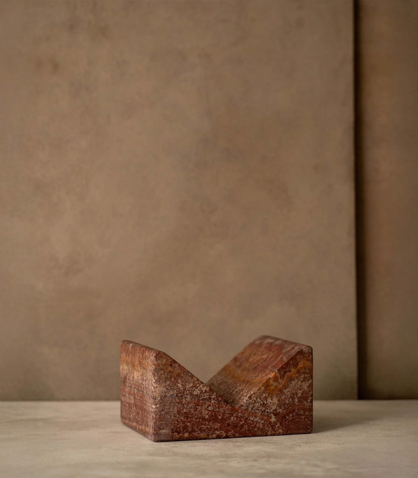 Close-up of a small, weathered wooden wedge with a rust-colored surface, placed on a light-colored surface against a plain, neutral-colored background.