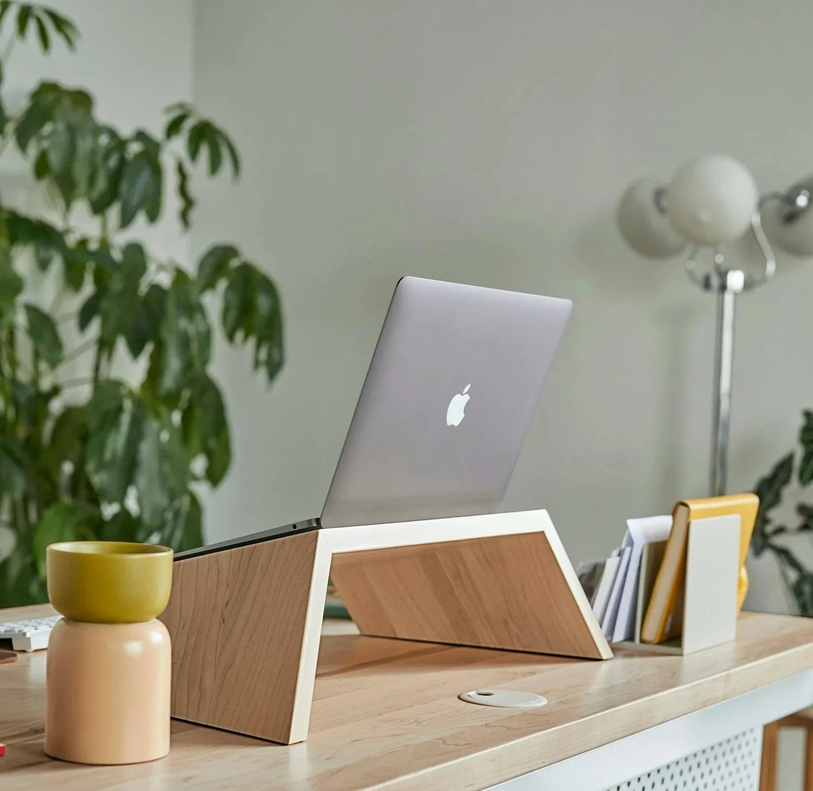 Wooden desk with a laptop, a yellow and beige ceramic cup, and a stack of books in a holder. Green plant and a wall with a modern lamp in the background.