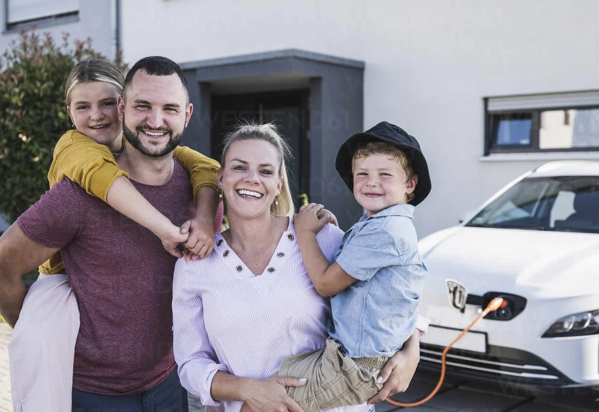 happy-family-standing-in-front-of-family-home-and-electric-car-UUF27087.jpeg