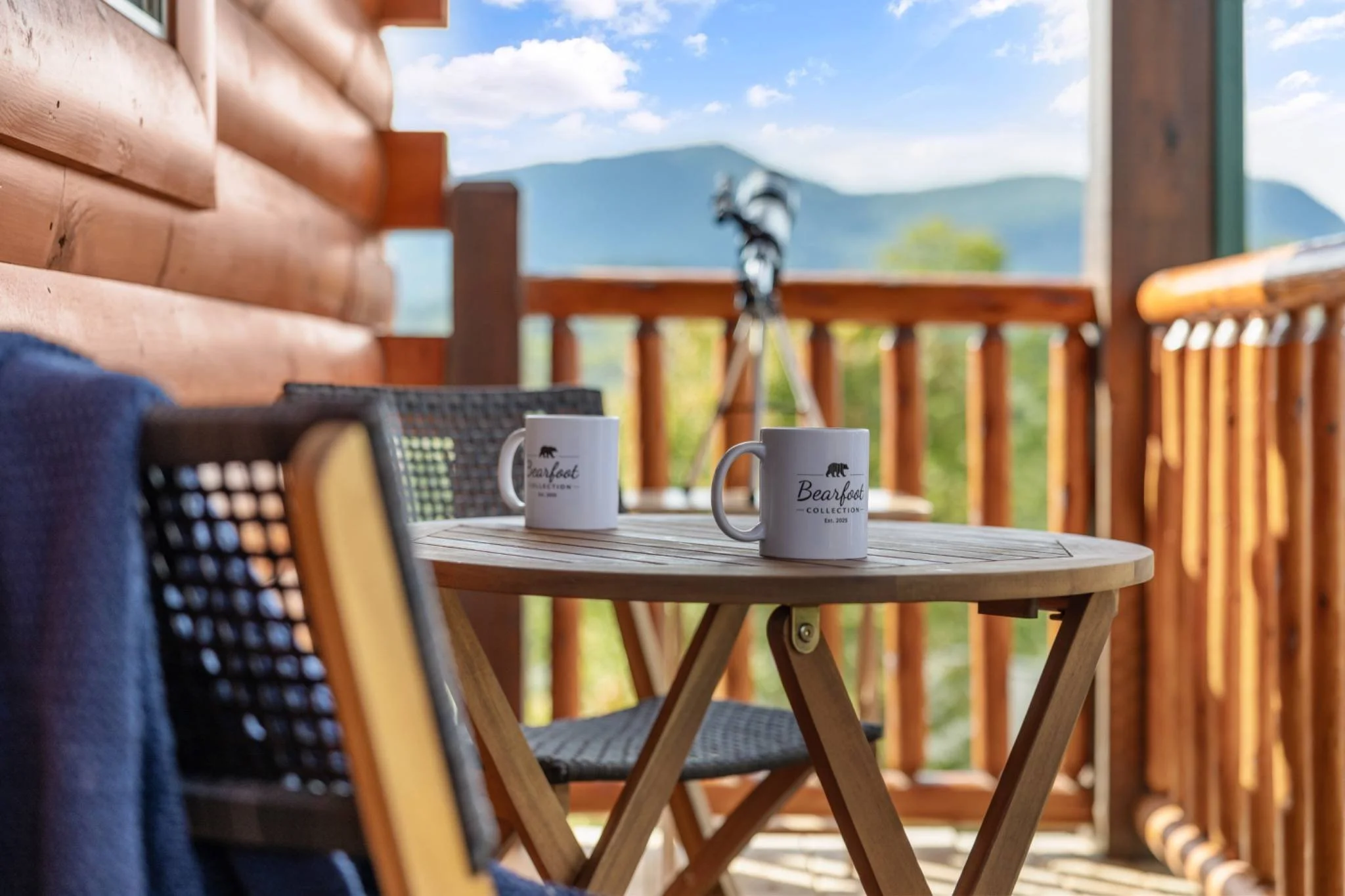 Cabin balcony with two Bearfoot Collection coffee mugs on a wooden table, mountain view in the background, Smoky Mountains rental.