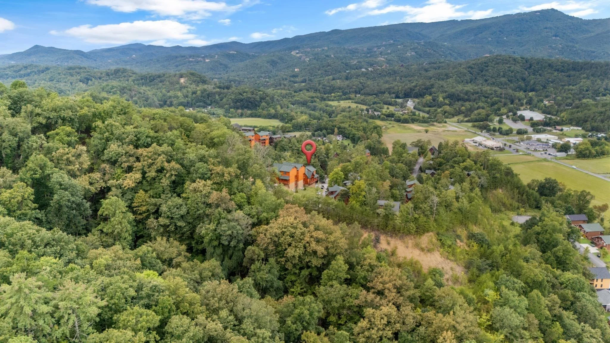 Aerial view of a wooded mountain landscape with a cluster of orange and gray buildings, a red location pin on one of the buildings, and a town with parking lots in the distance under a partly cloudy sky.
