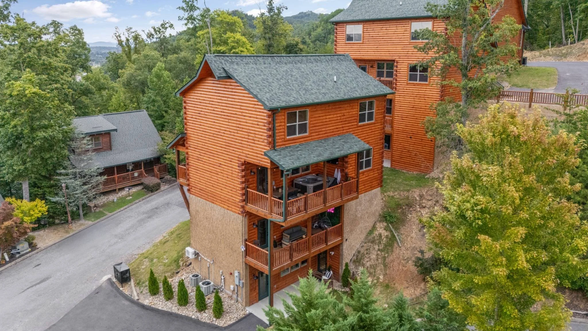 An aerial view of a multi-story log cabin house with green shingle roof, surrounded by trees and other cabins, in a wooded area.
