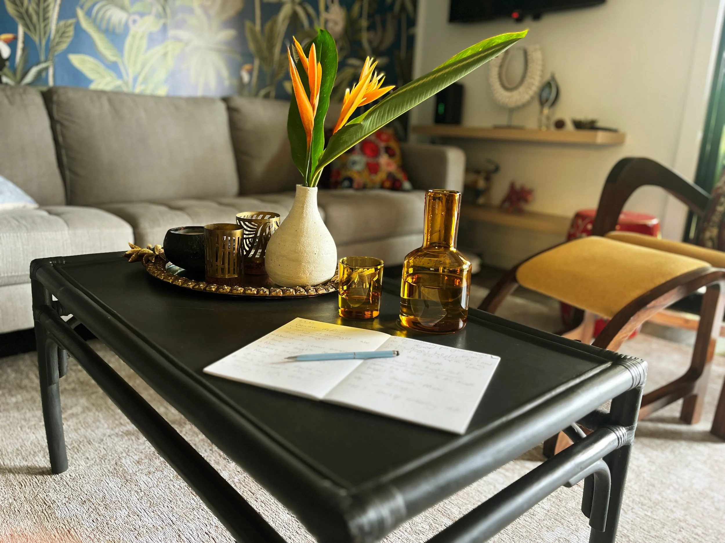 Living room with a black coffee table holding a notepad with writing and a pen, a ceramic vase with tropical leaves and orange flowers, amber glass pitcher, glass, and decorative items. In the background, there is a beige sofa, a patterned wall, and other furniture.