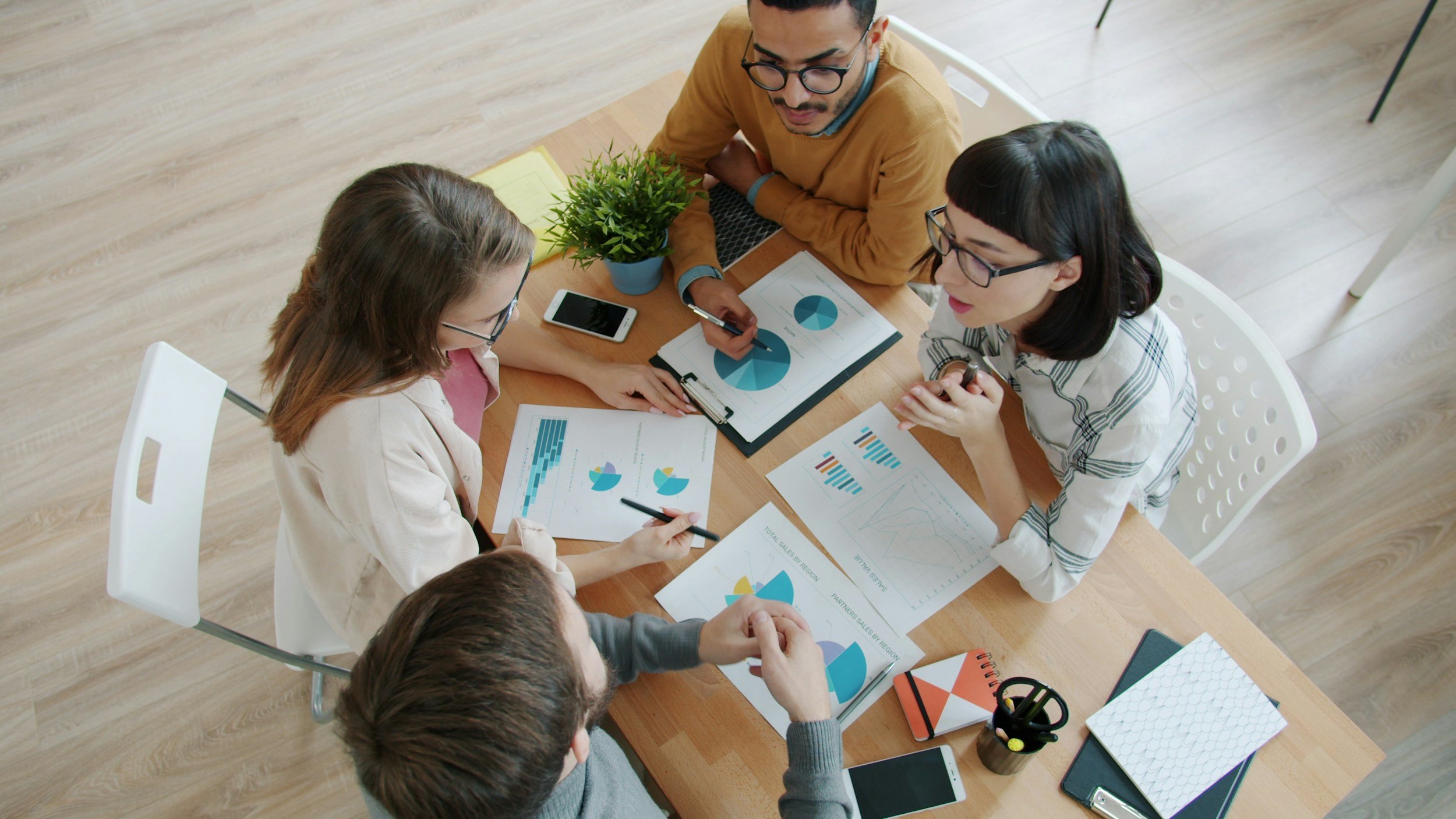 A group of five people in a meeting room discussing charts and graphs on papers and a tablet, with a plant on the table.