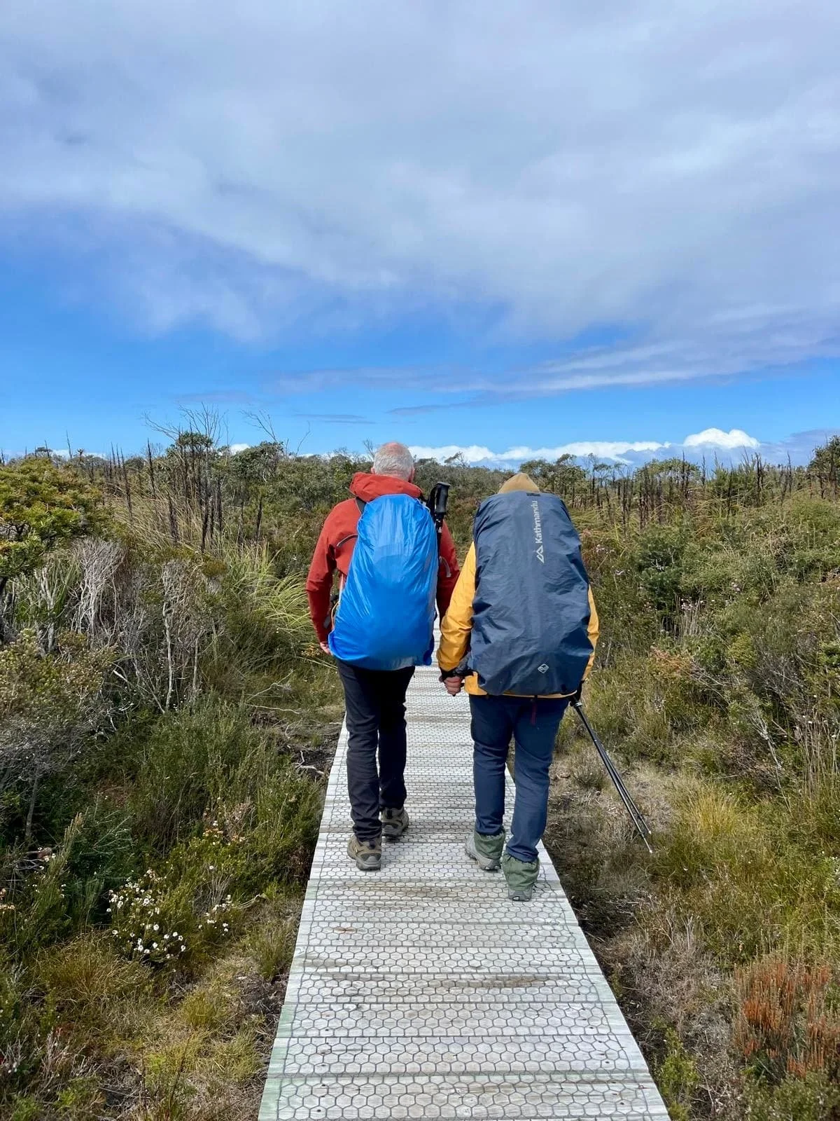 Joanna and Bruce walking hand-in-hand along the 3 Capes hiking trail in Tasmania, surrounded by native grasses and coastal bushland under a soft blue sky.
