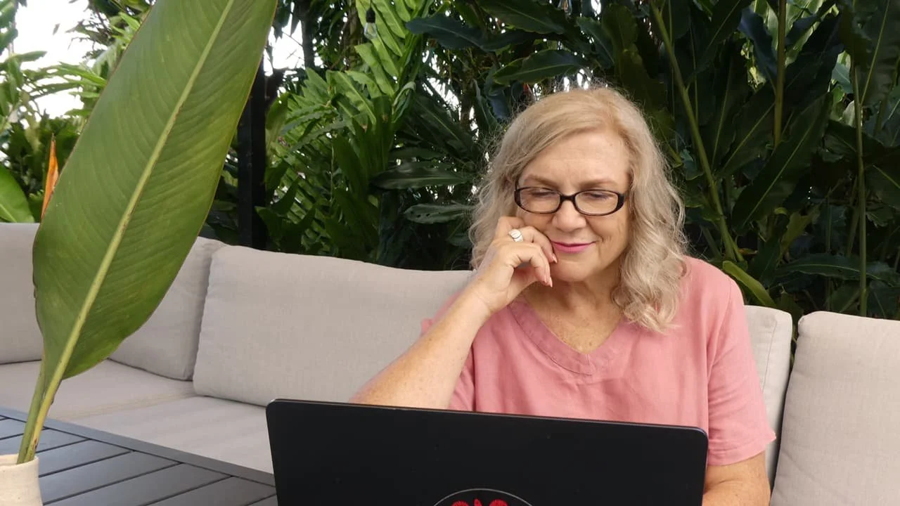 A woman with blonde curly hair and glasses working on a laptop, sitting on an outdoor sofa surrounded by lush green plants.
