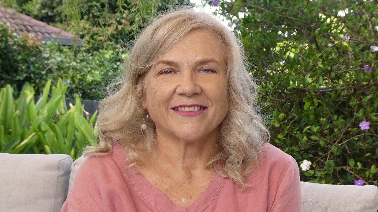 Joanna Wood Counsellor for midlife women sitting outdoors on a white couch, with green plants and purple flowers in the background.