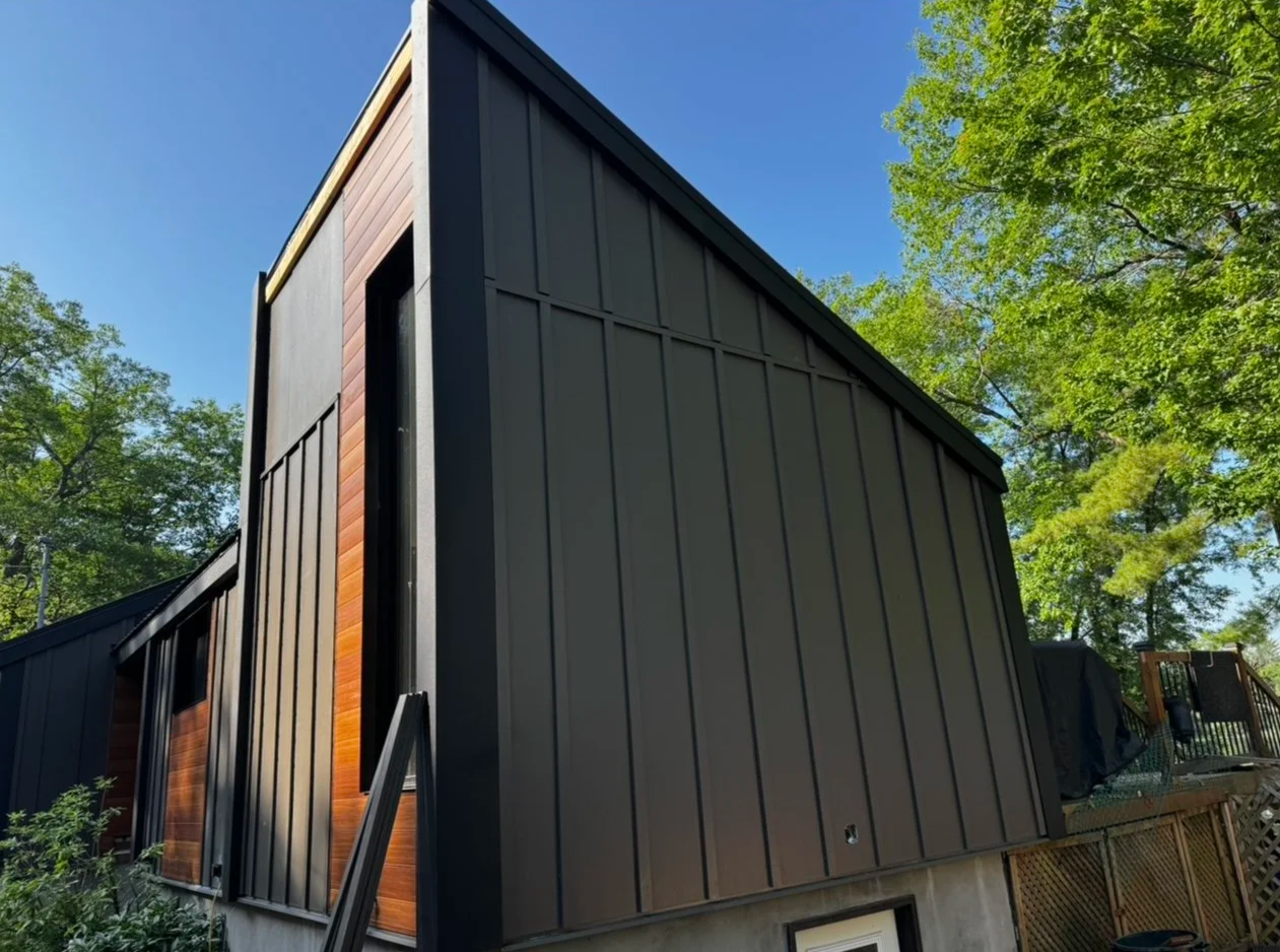 Side view of a modern black and wood-paneled house with a sloped roof, surrounded by green trees and a clear blue sky.