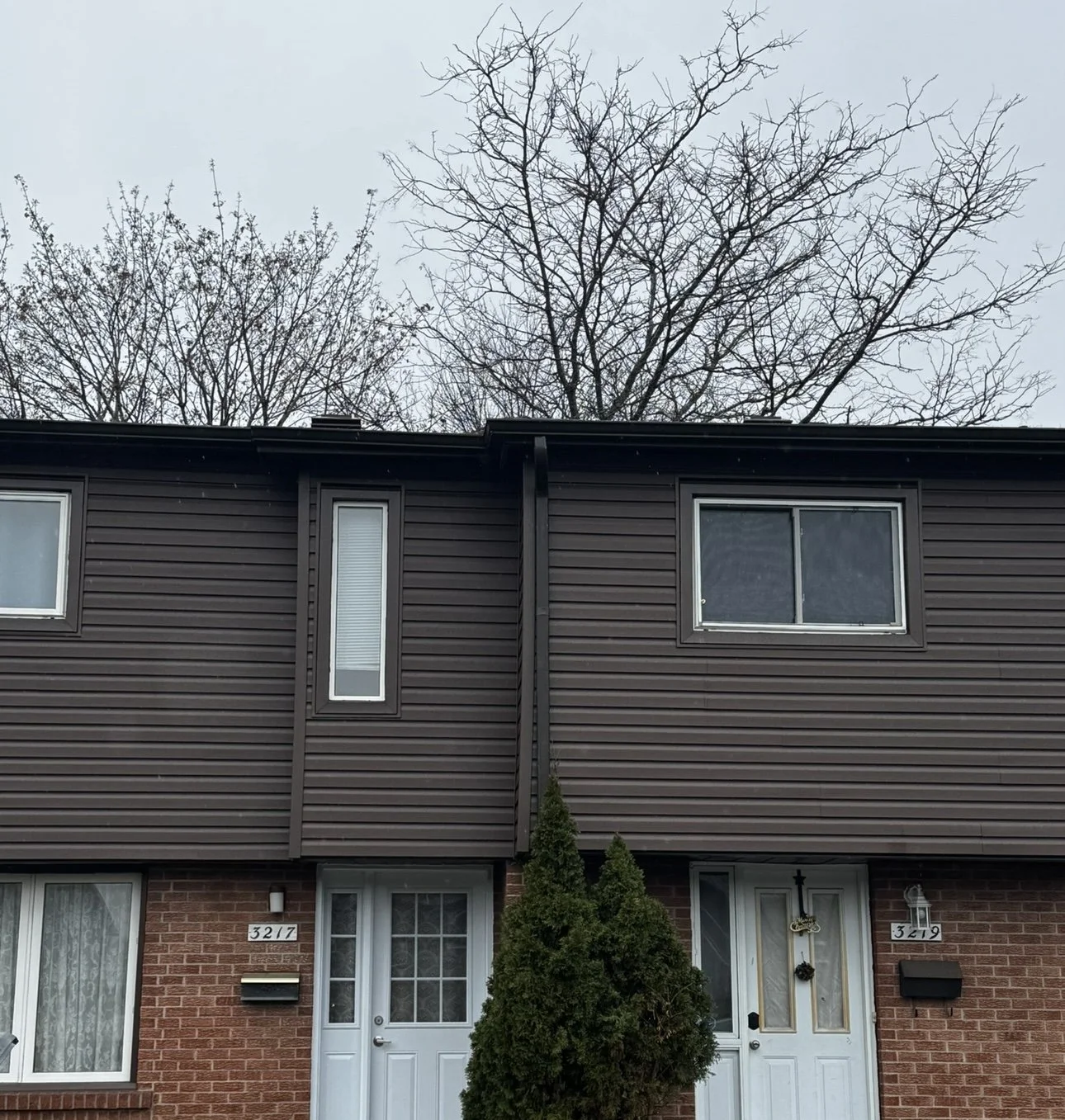 Two adjacent brick townhouses with brown siding on upper floors, each with a white front door, small window, and black mailbox. Two trees with no leaves stand behind the houses against a gray, cloudy sky.