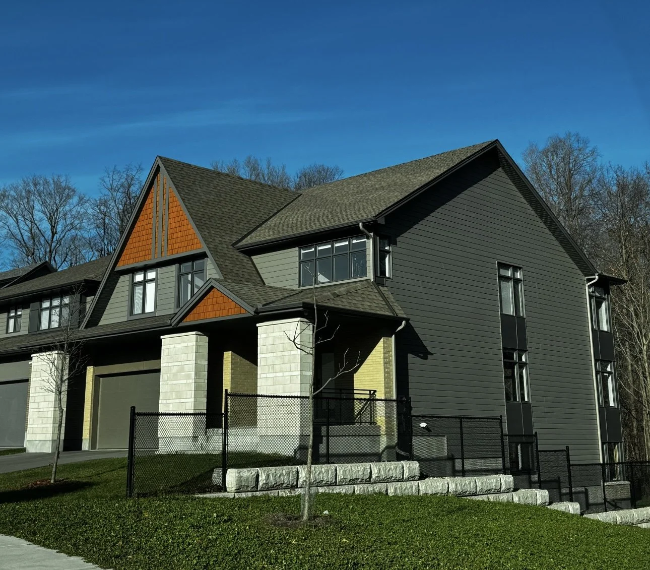 Modern multi-story house with black siding, large windows, and a mix of sloped roof styles, surrounded by a small yard with a young tree and a black fence.