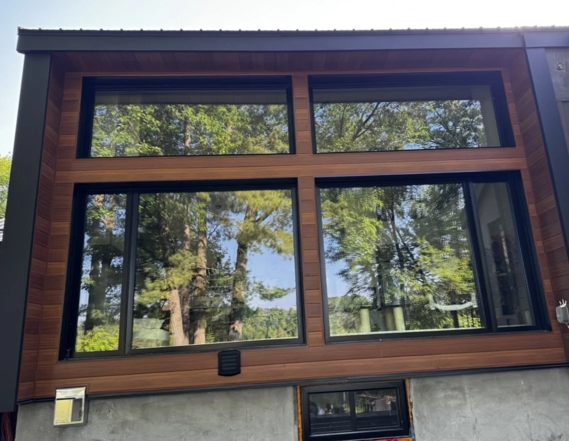 The exterior of a modern house with large glass windows and wooden framing, reflecting trees and sky.