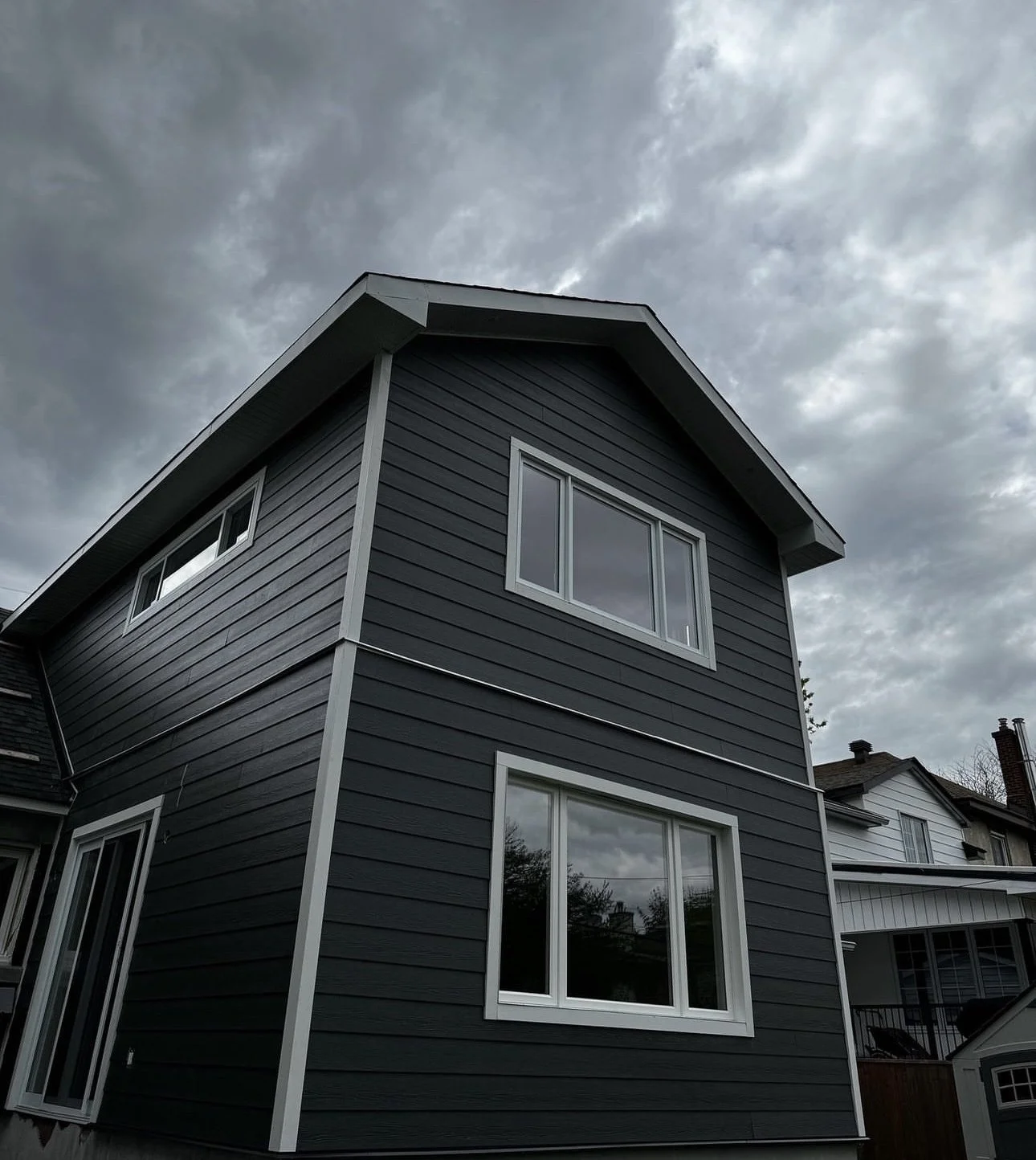 A modern two-story house with dark gray siding and white window frames under a cloudy sky.