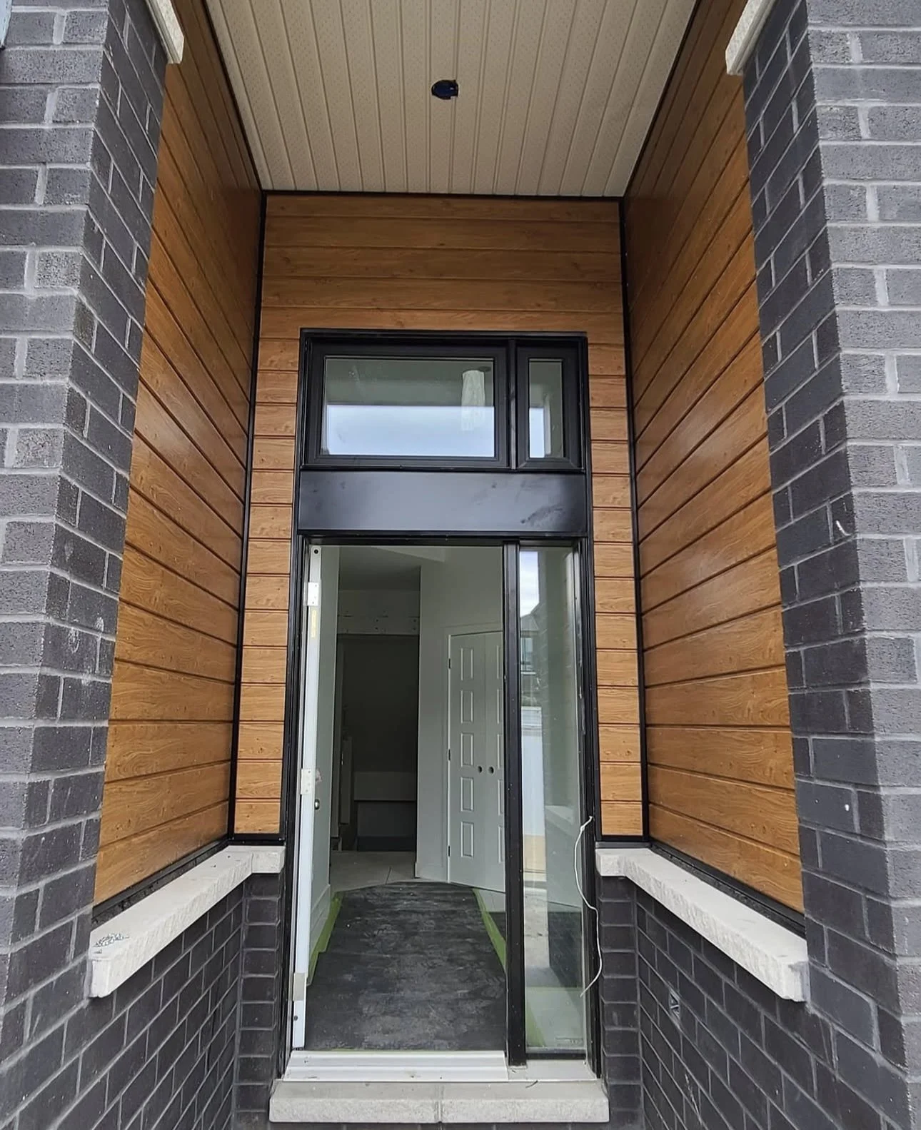 Close-up view of a residential entryway with an open glass sliding door, black window frames, and exterior siding made of wood panels surrounded by brick walls.