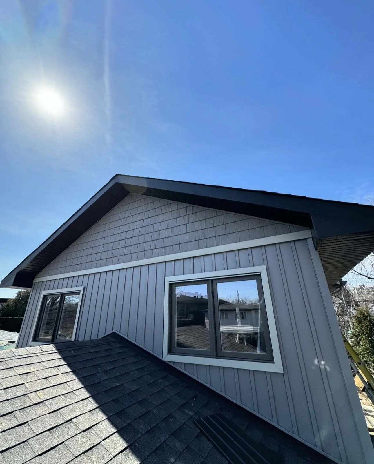 View of a modern house with gray siding and a gable roof, featuring a large window with a panoramic view, under a clear blue sky with the sun shining brightly.