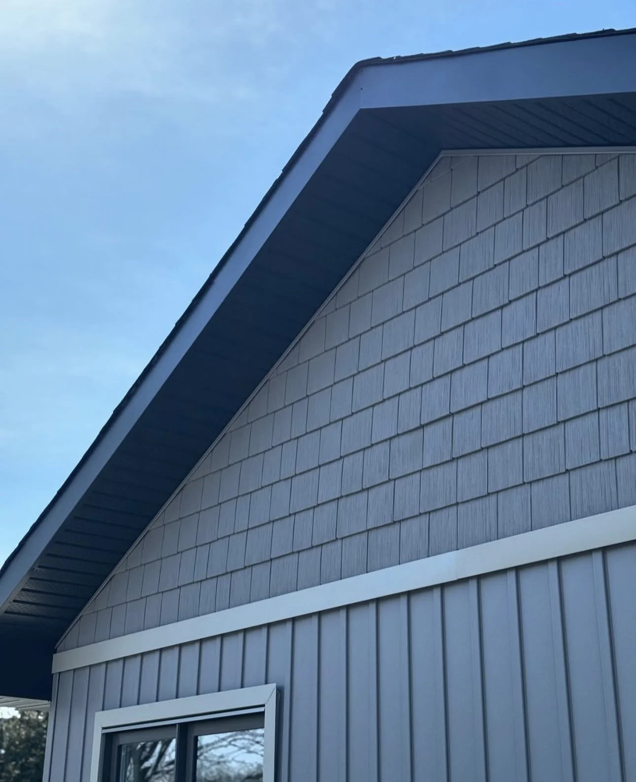 Close-up of the side of a modern house with gray vinyl siding, a roof overhang, and a window, against a blue sky.