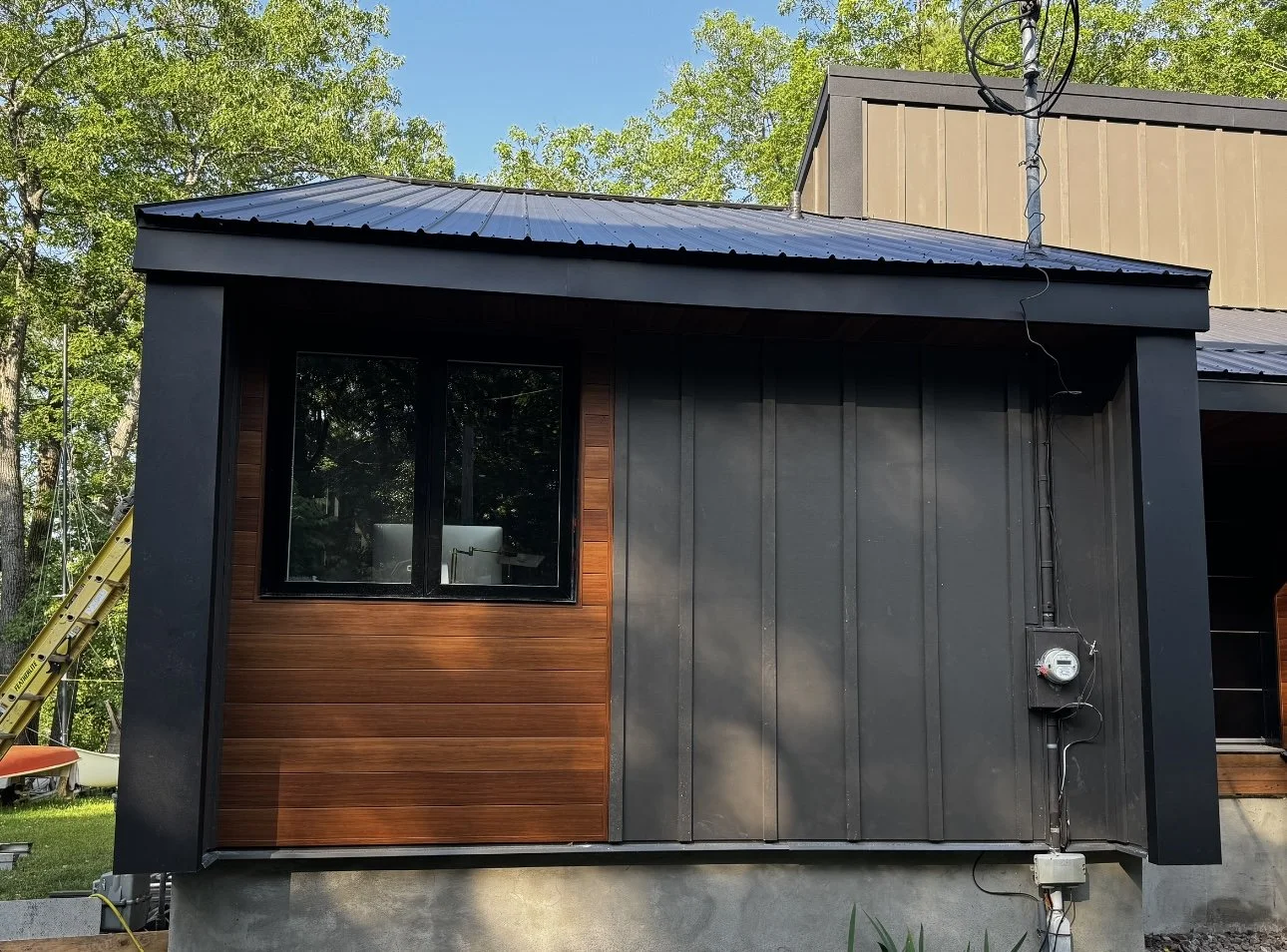 Exterior view of a modern house with a black metal roof, a large window, and black and wooden wall panels, surrounded by green trees.