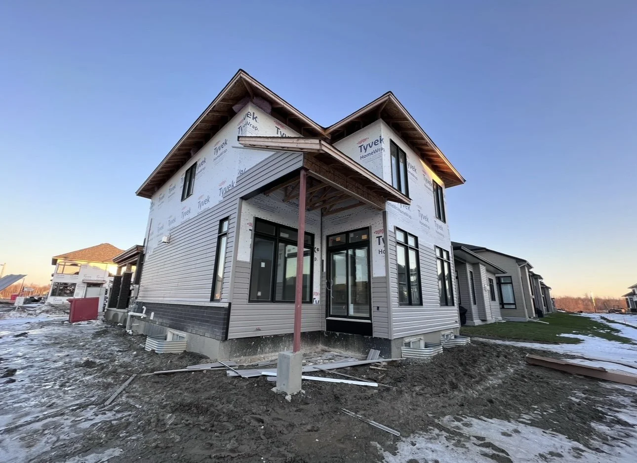 A two-story house under construction with siding and large windows, surrounded by bare ground and snow on a clear evening.