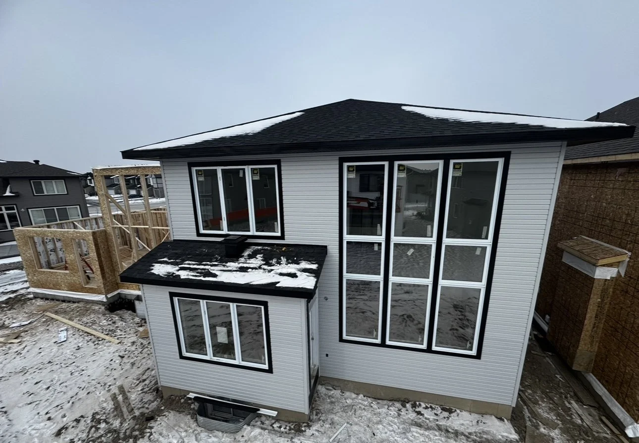 Two-story house under construction with siding, large windows, and a black roof, surrounded by snow and outdoor building materials.