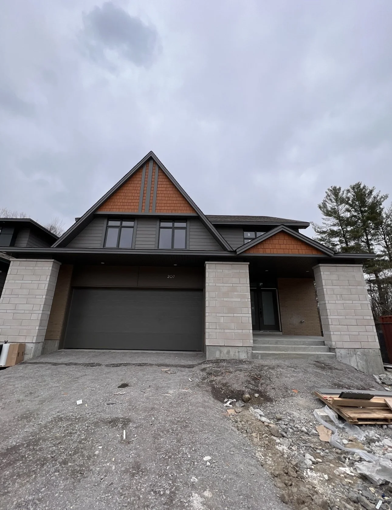 Front view of a modern house under construction with a driveway, garage, and steps leading to the front door, overcast sky, and trees in the background.