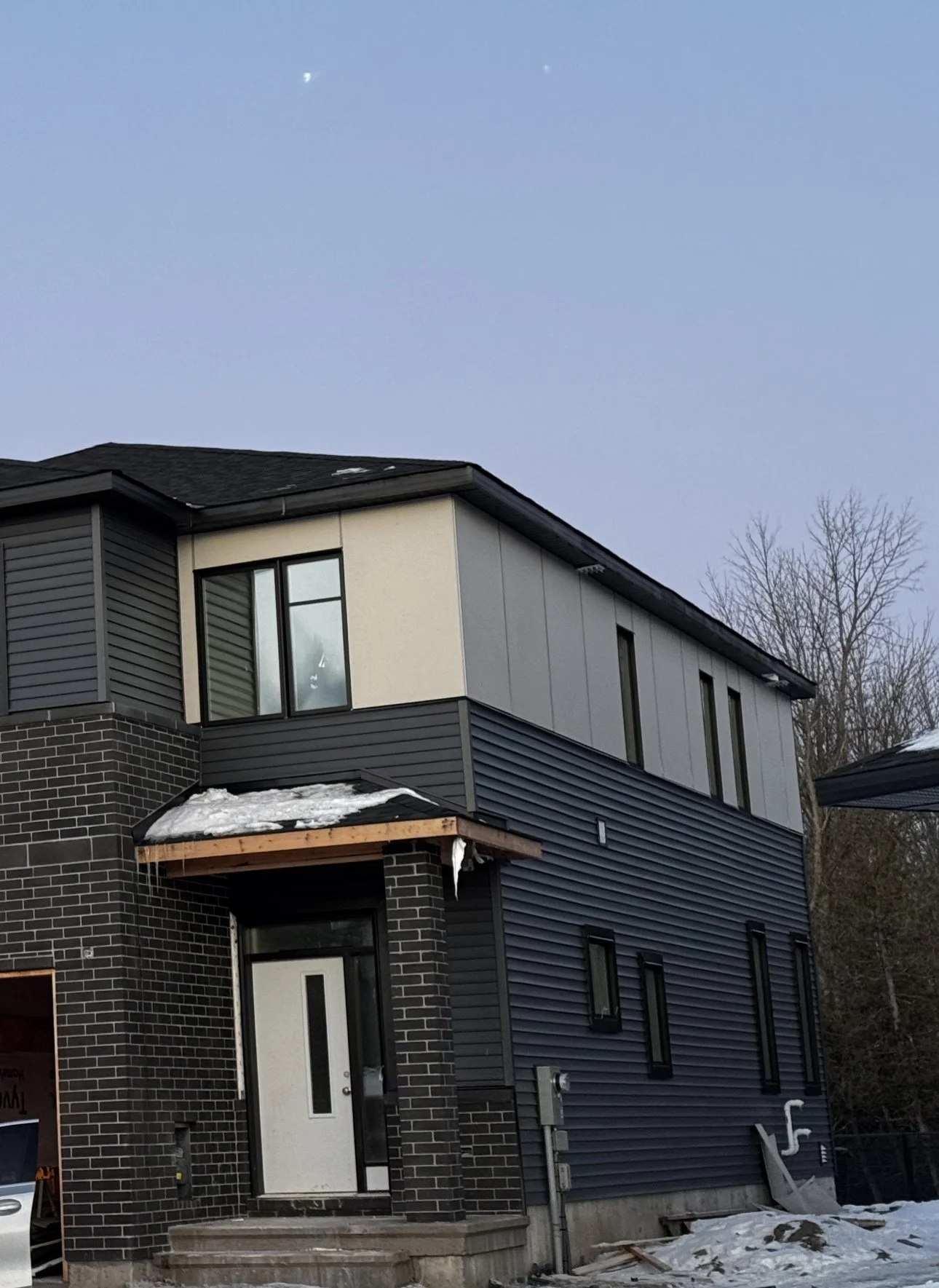 Modern multi-story house with black brick and gray siding exterior, some snow on the roof, and a front door with steps, during winter.
