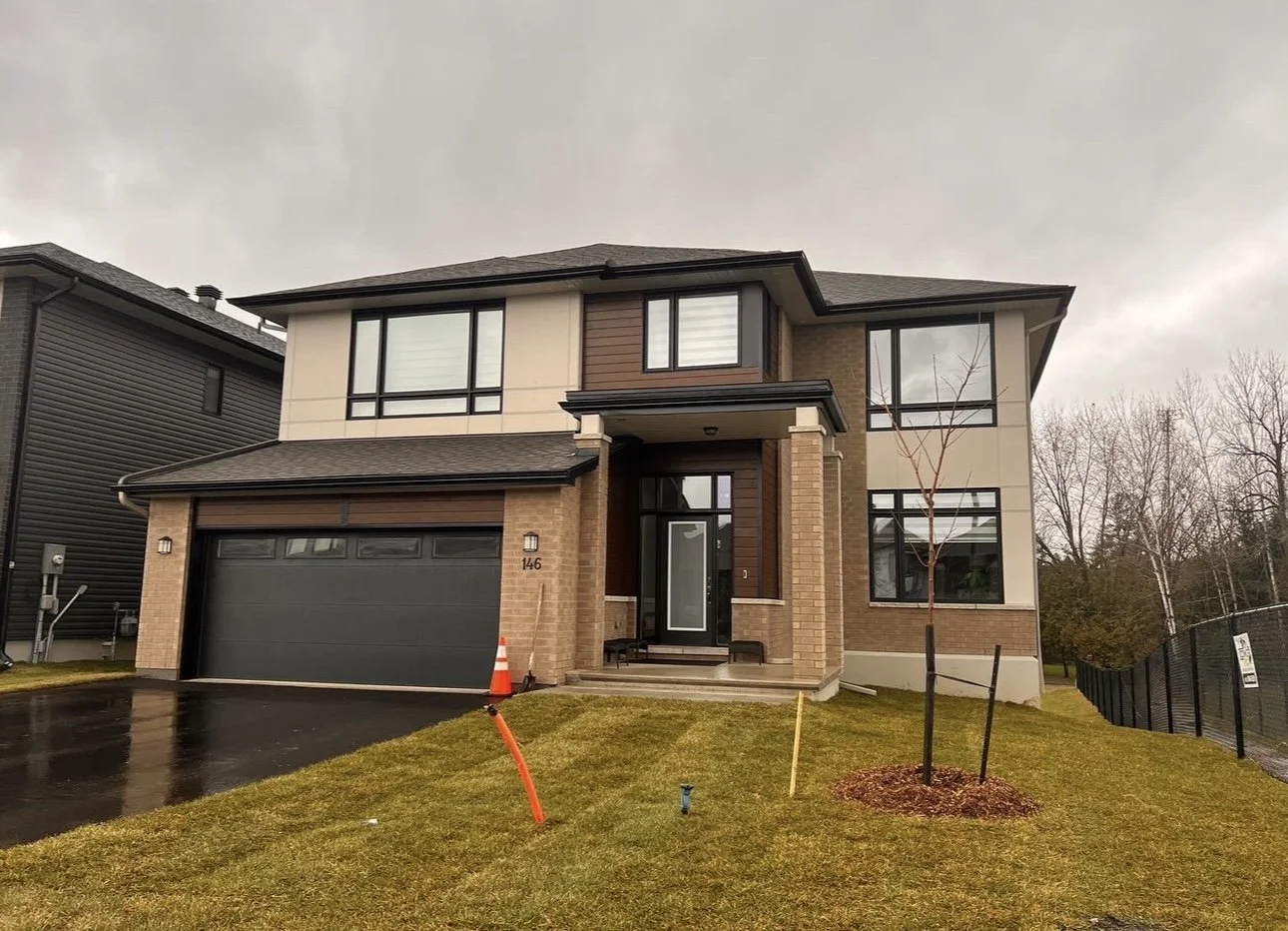 Newly built modern two-story house with brick and siding exterior, large windows, and a dark garage door, with a small tree in front yard and overcast sky.