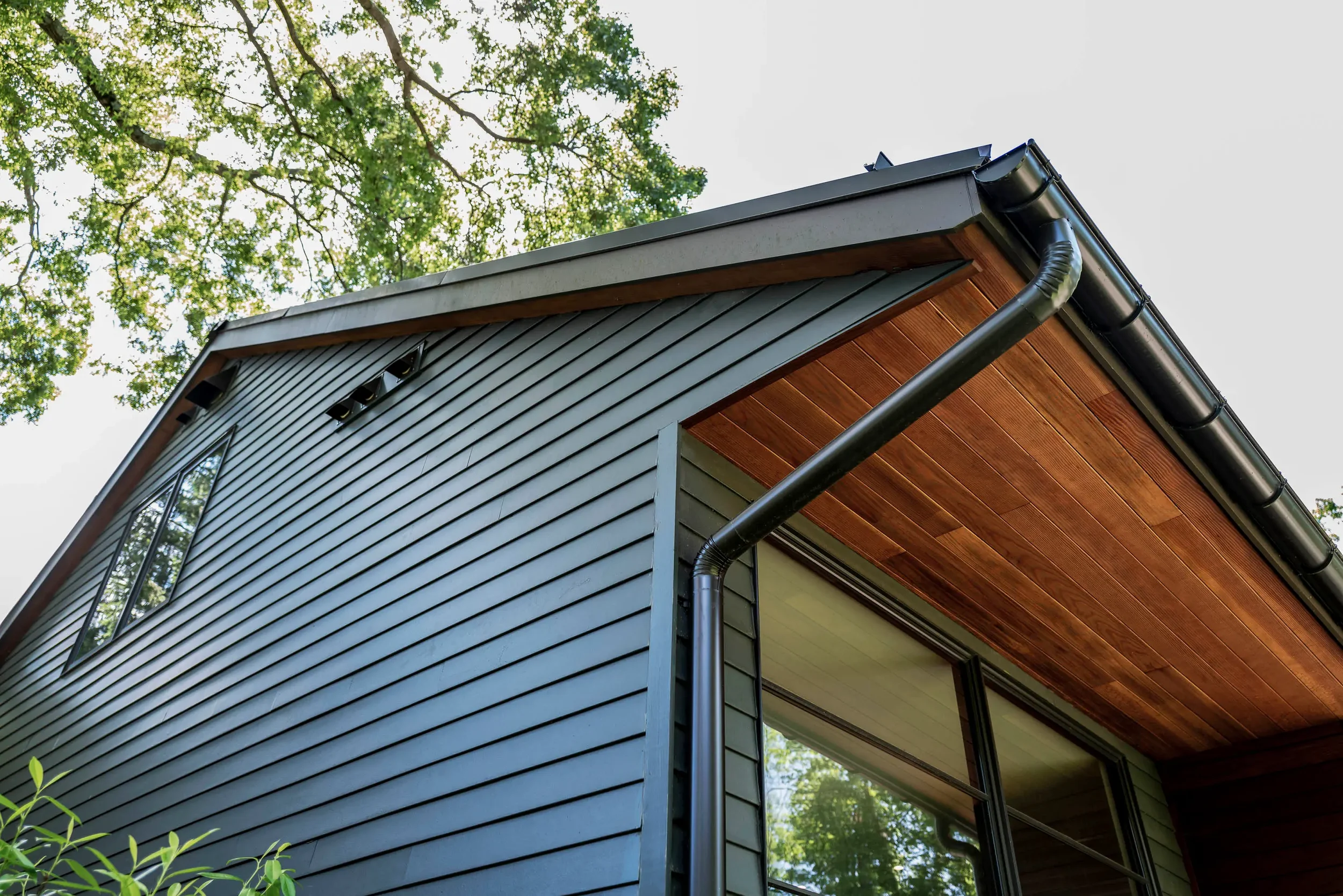 Close-up of the corner of a modern house exterior showing dark horizontal siding, a rain gutter, and a wooden ceiling under the roof overhang, with trees in the background.