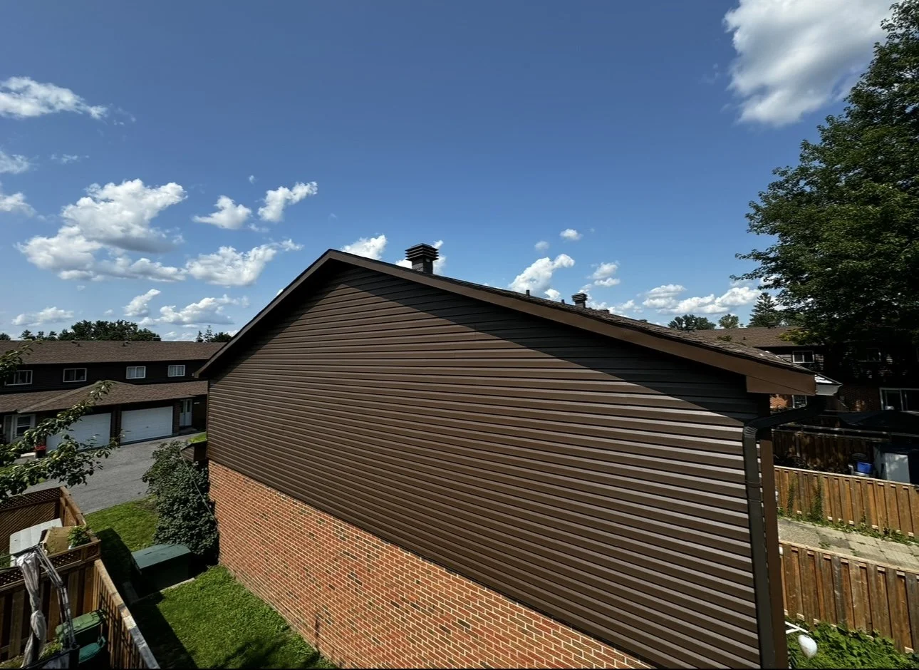View of the side of a house with a sloped roof, brown siding, brick lower wall, and a backyard with a wooden fence, trees, and a bright blue sky with some white clouds.