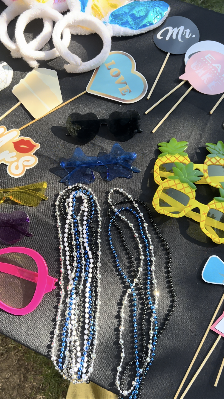 A collection of colorful party props on a black table, including silly glasses, necklaces, small signs with words like "LOVE" and "Mr.", white fluffy headbands, and pineapple-shaped sunglasses.
