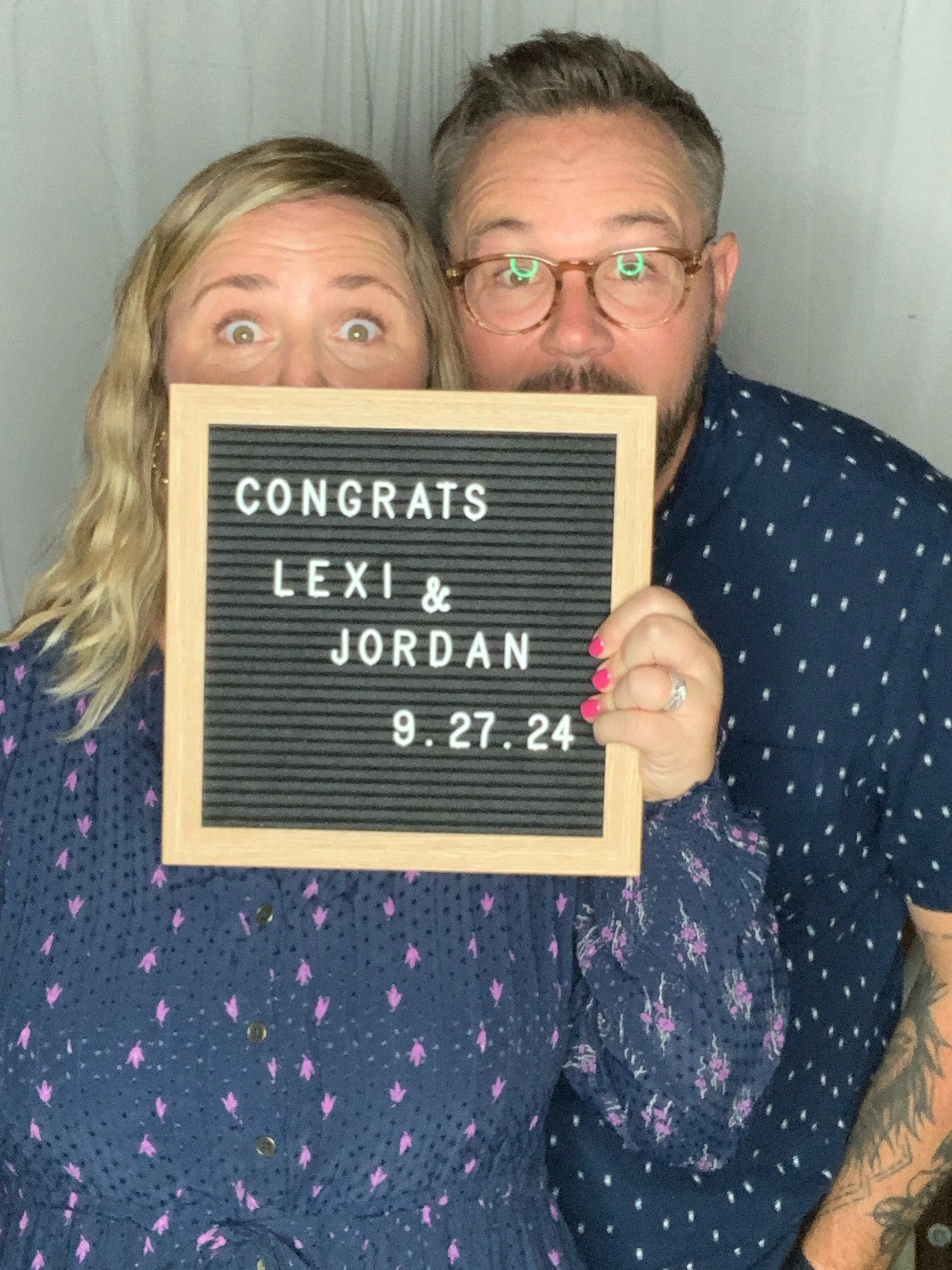 A man and woman posing with a letter board that says, 'Congrats Lexi & Jordan 9.27.24', holding it close to their faces, both looking at the camera with wide eyes, in front of a gray wall.