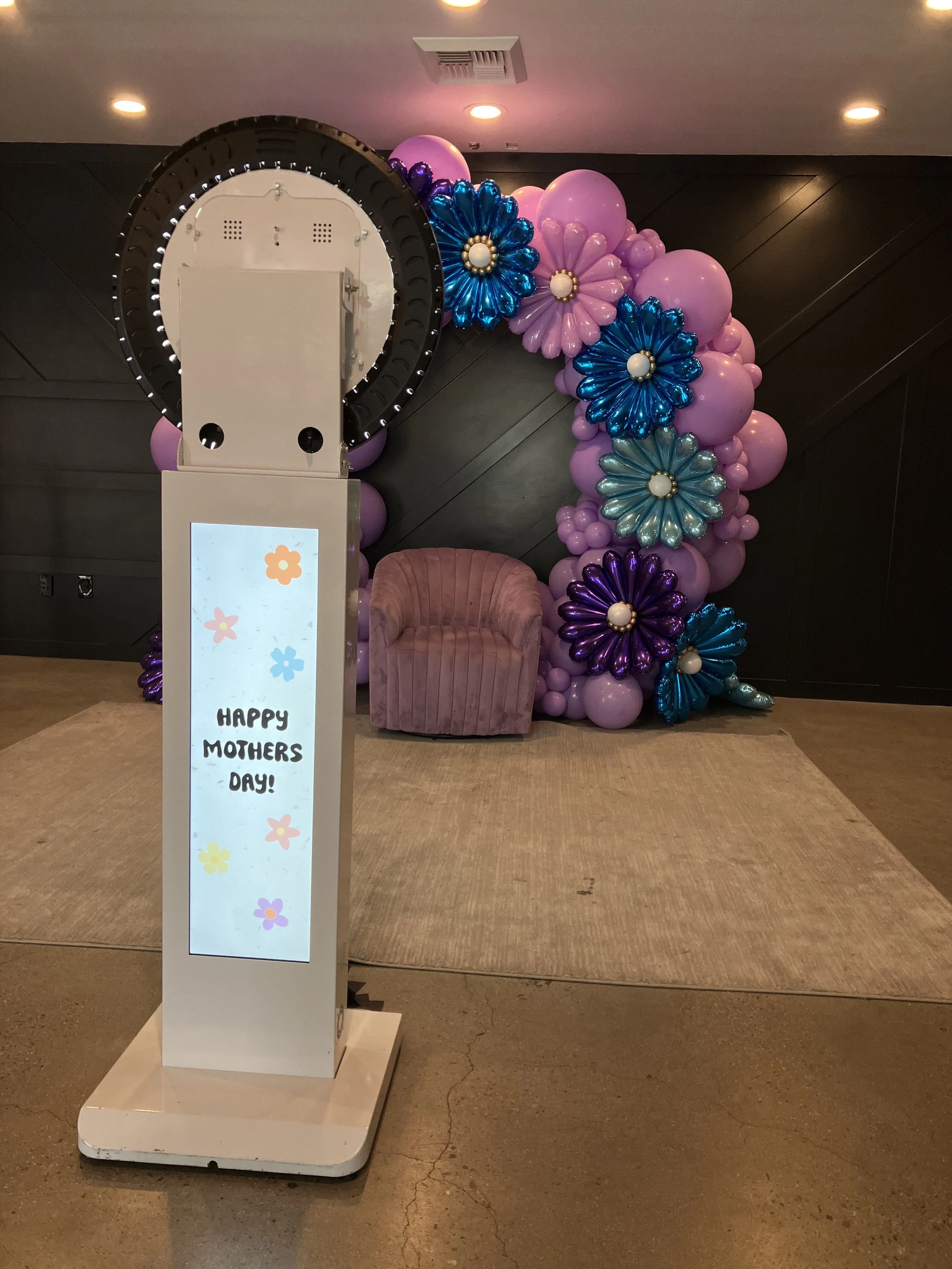 Photo of a photo booth with a floral balloon backdrop and a pink chair, with a digital screen displaying a message that says 'Happy Mothers Day!'