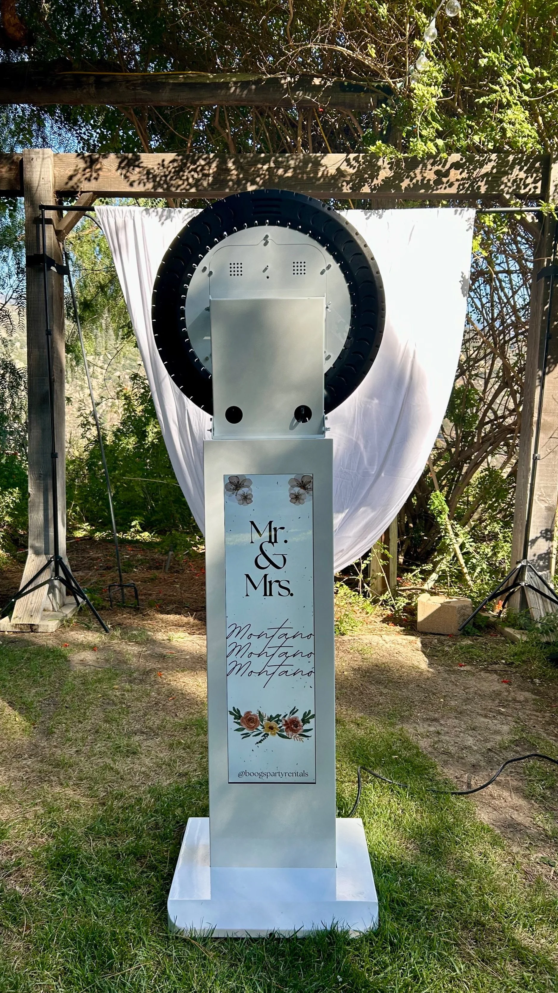 A photo booth with a circular photo capture ring and a sign that says 'Mr. & Mrs.', adorned with floral graphics, set outdoors with a white backdrop and greenery.