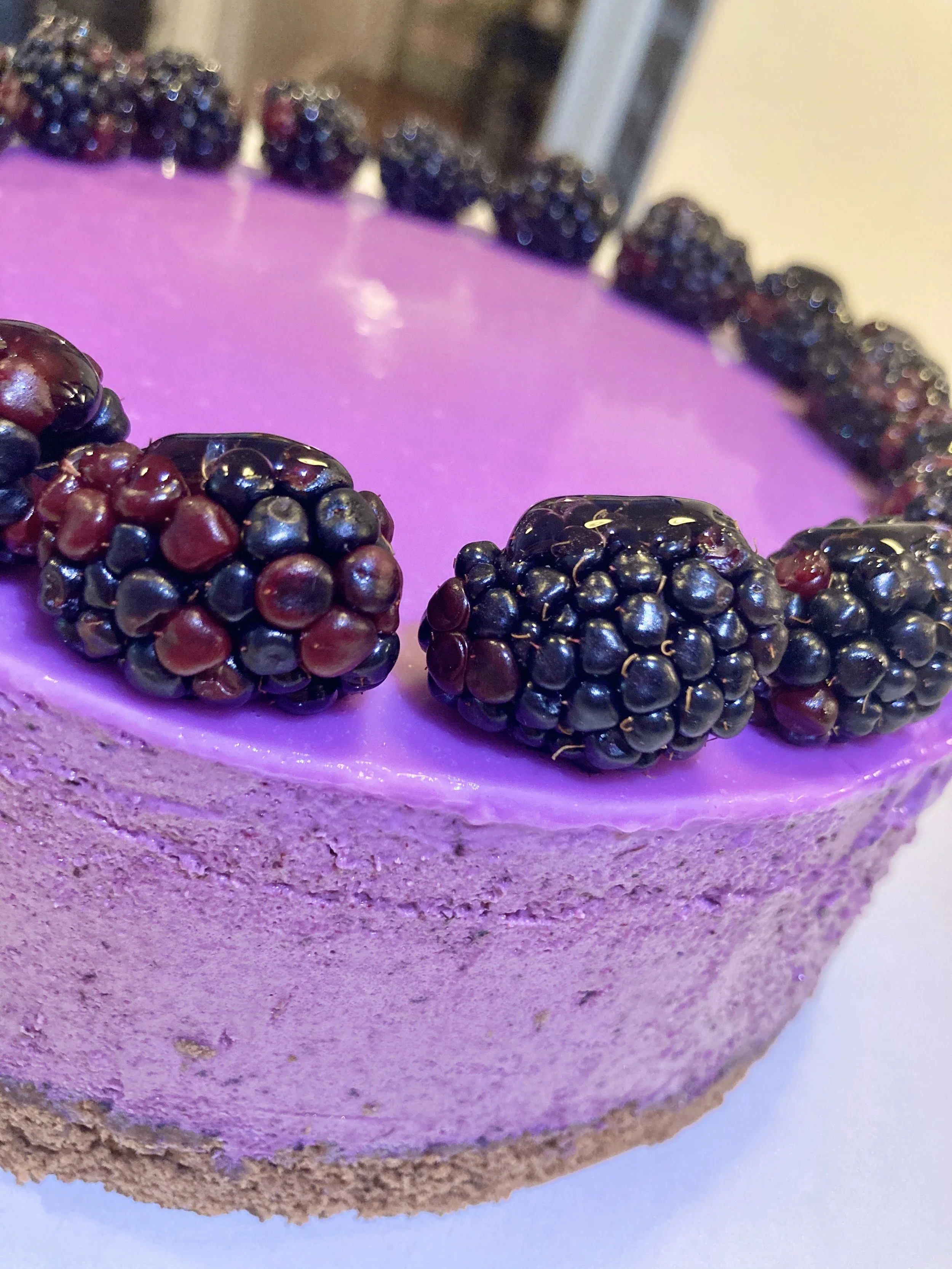 A close-up of a purple blackberry-flavored cheesecake with a graham cracker crust, decorated with fresh blackberries around the top edge.
