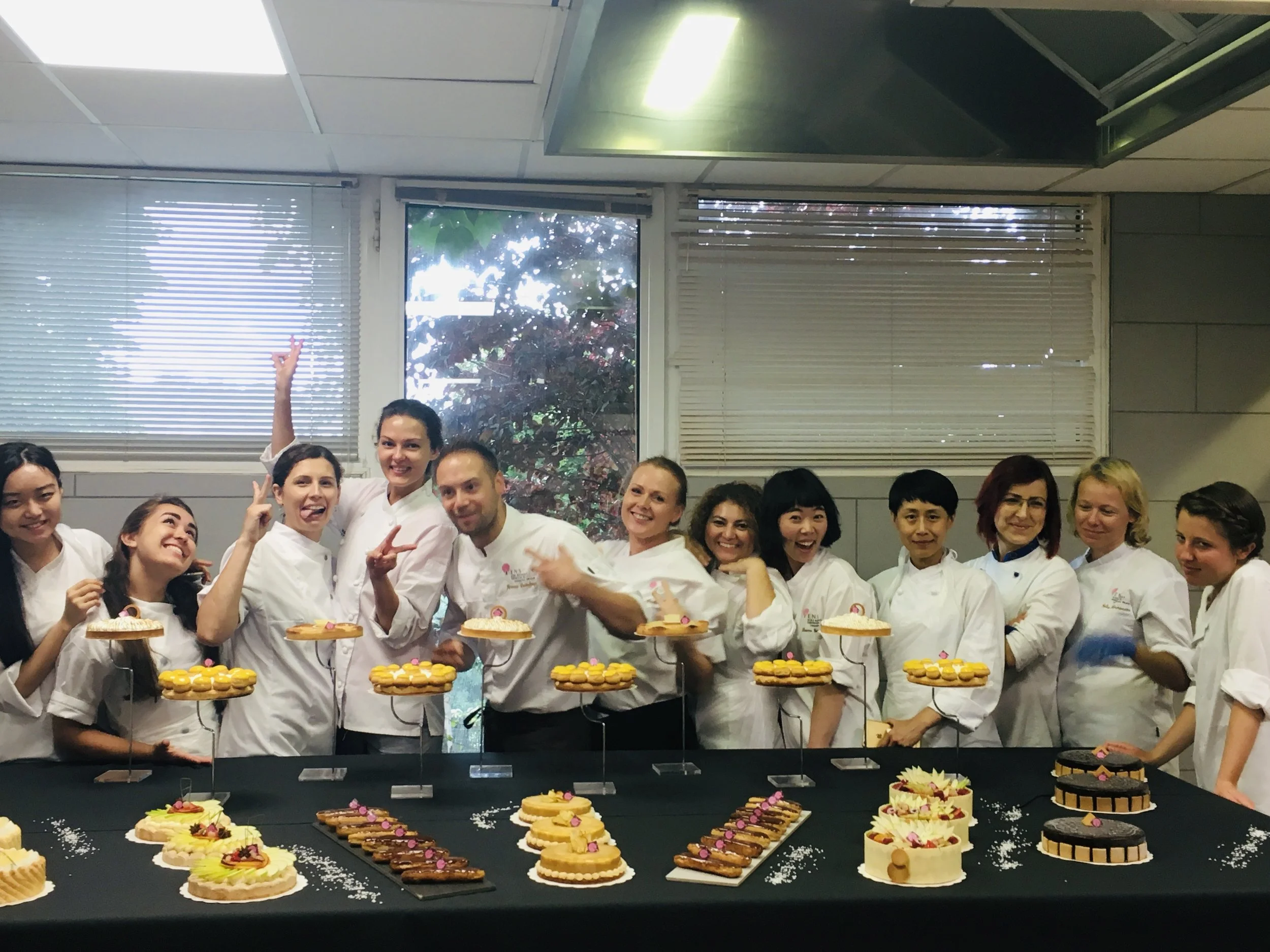 Group of pastry chefs and bakers celebrating behind a table of decorated cakes and desserts.